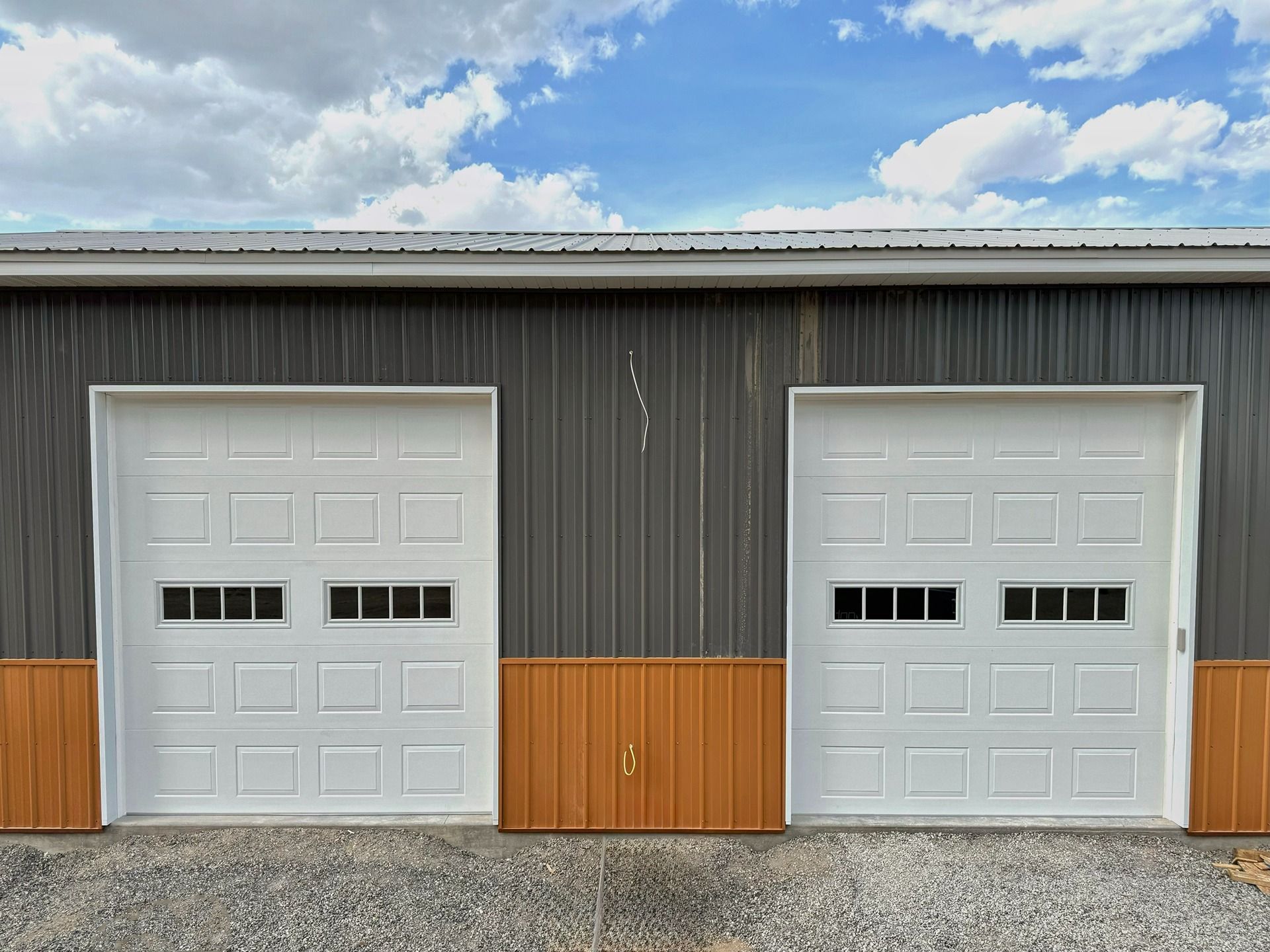 Two white garage doors with small windows set into a dark gray and orange metal building under a blue sky with clouds.