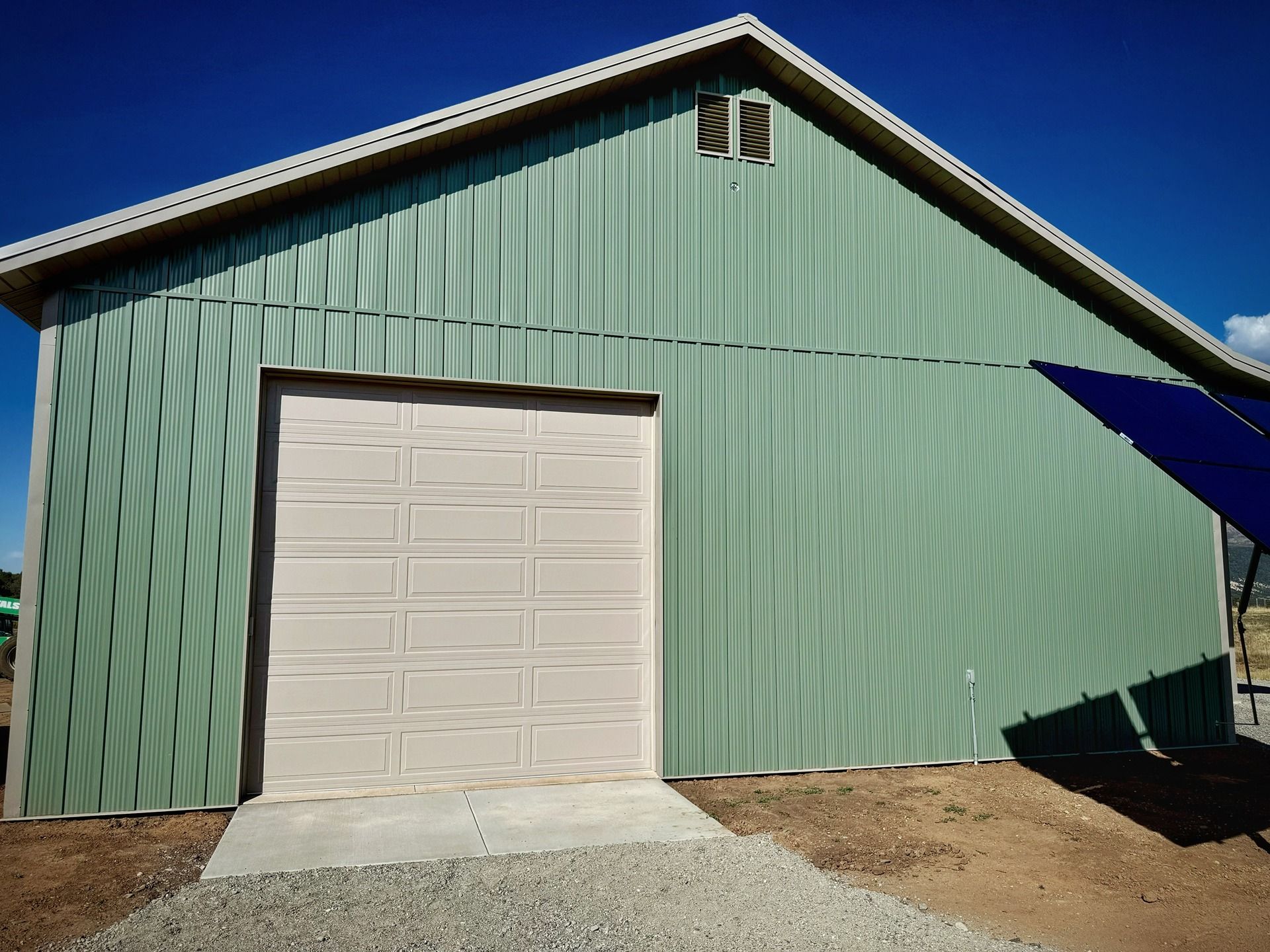A light green metal barn with a beige garage door and a gravel driveway under a clear blue sky.