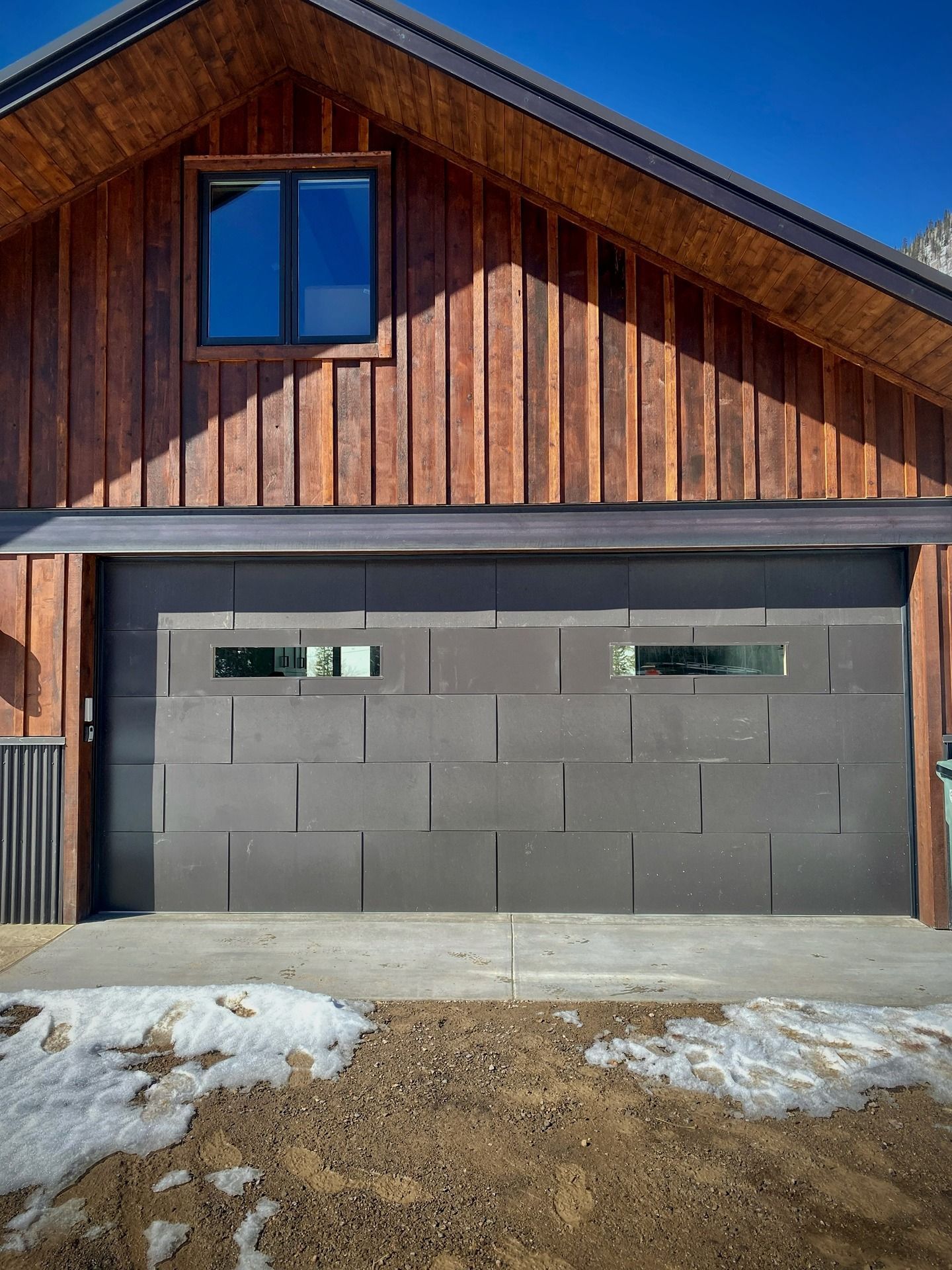 A dark, rectangular garage door with two narrow, horizontal windows, set into a rustic wooden barn-style building.