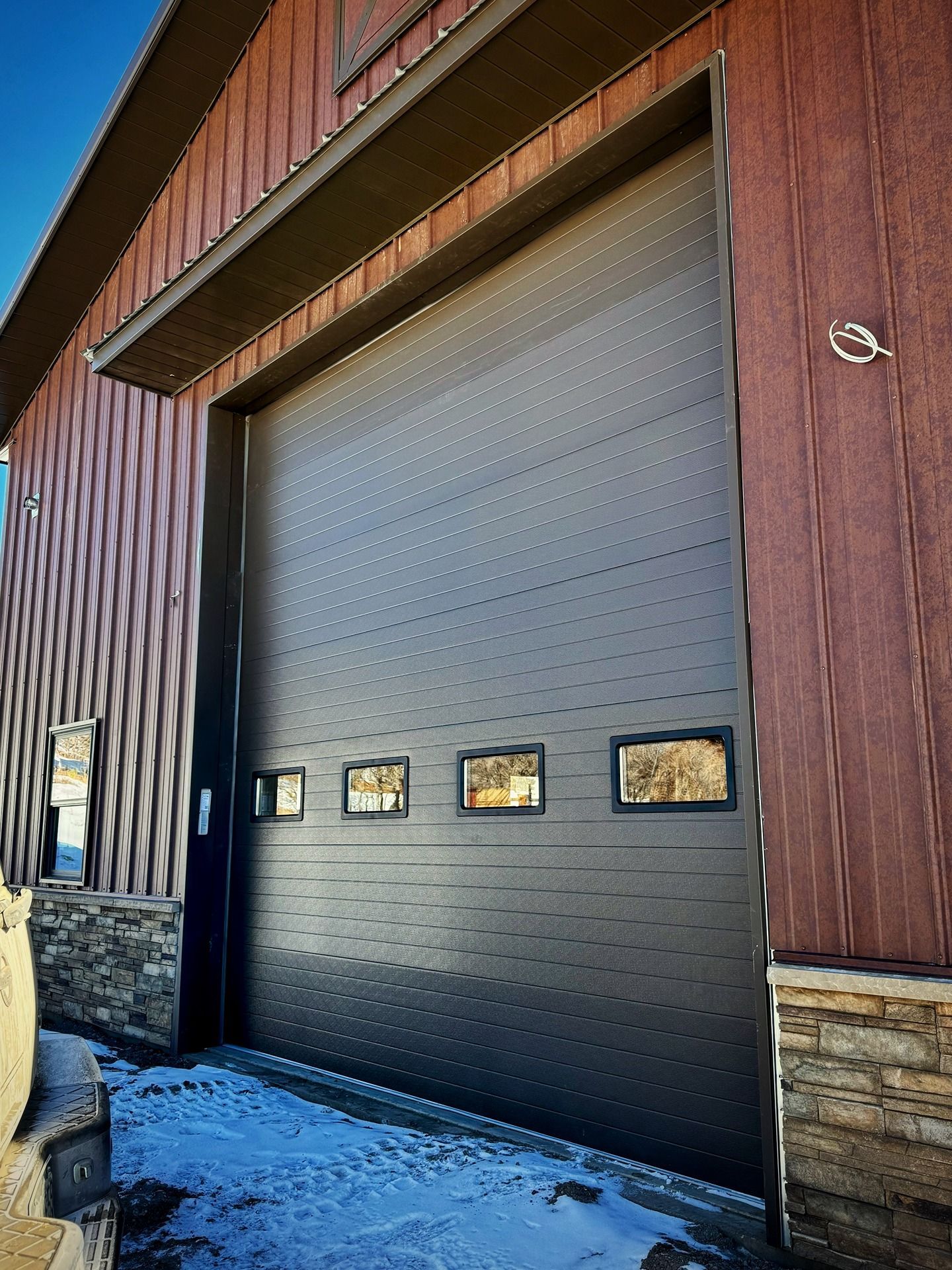 A large, dark brown garage door with four small windows, set in a building with wood siding and stone trim.