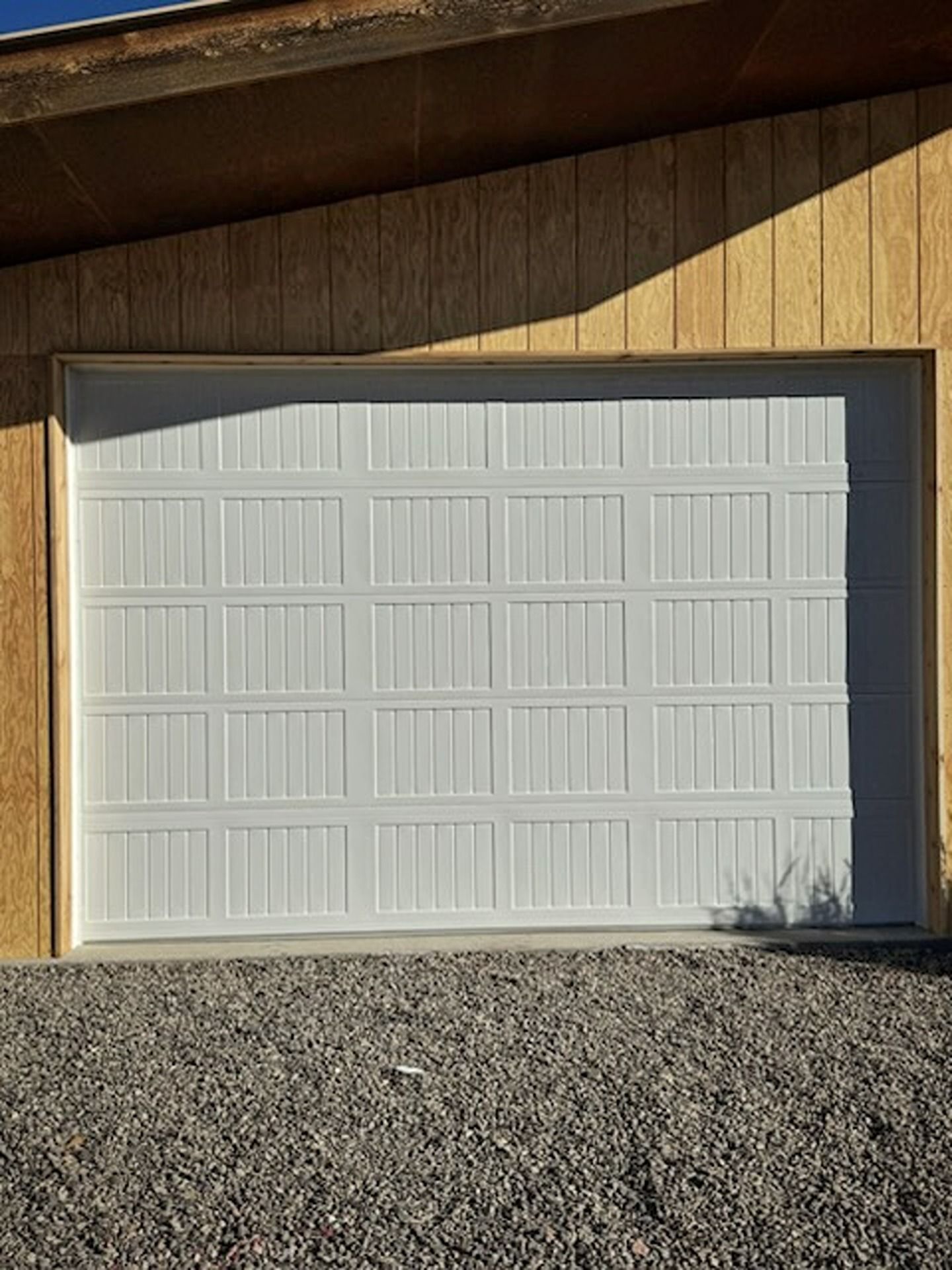 A white, multi-paneled garage door framed in light wood on a gravel surface.