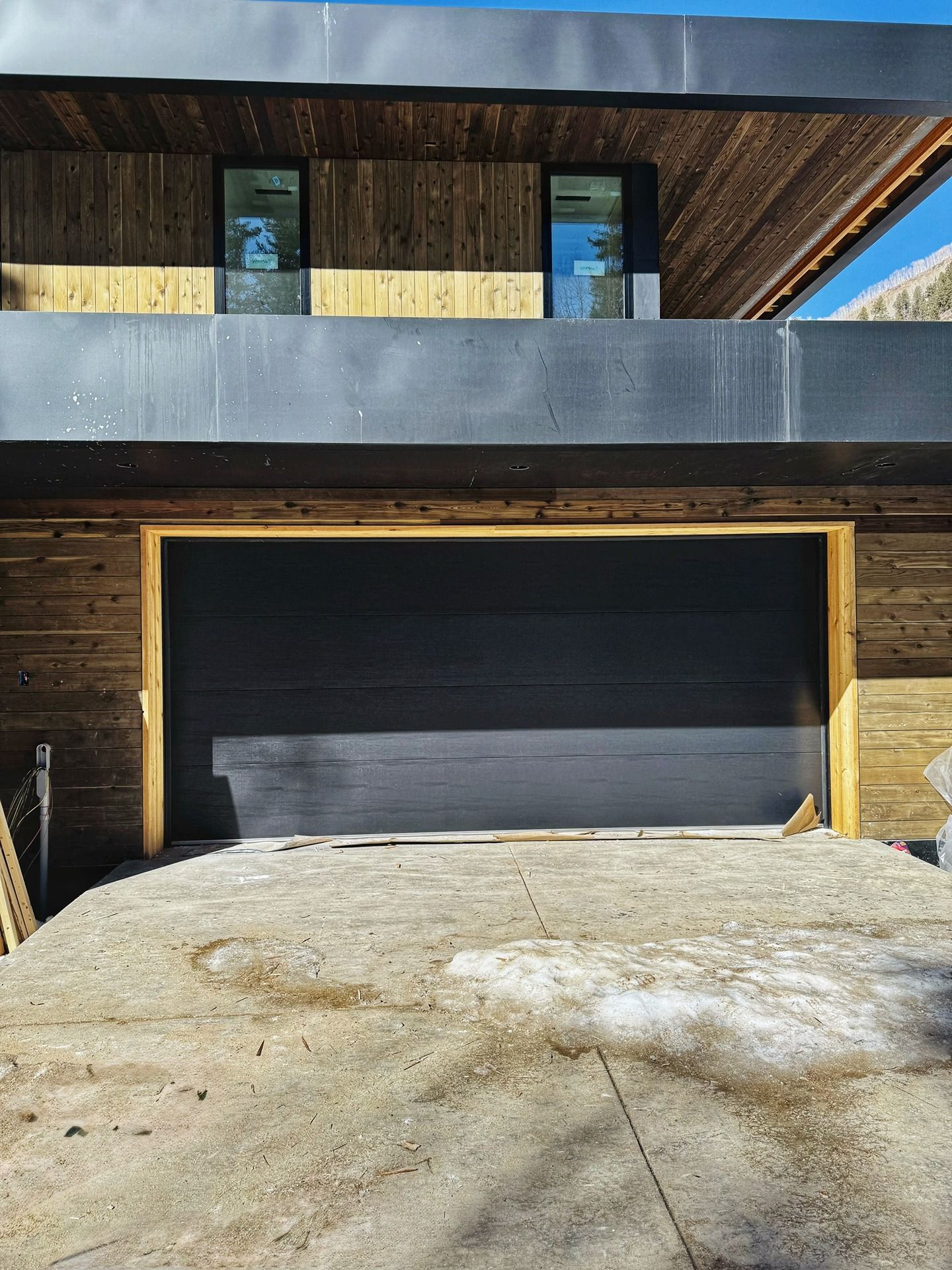 Modern house with wooden siding, a dark metal roof overhang, and a large, black horizontal-panel garage door.