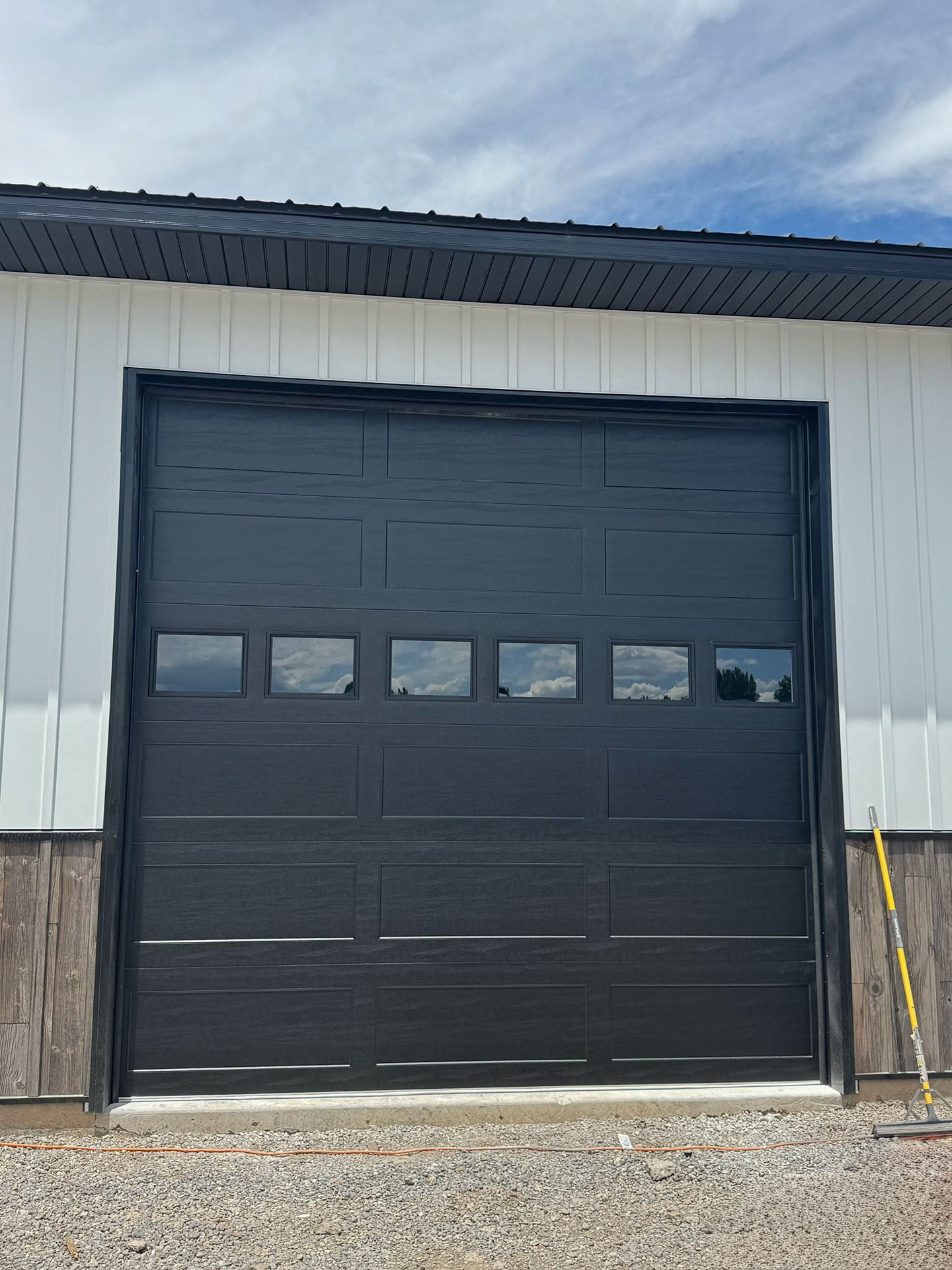 Black sectional garage door with a row of six rectangular windows on a white building exterior with a gravel driveway.