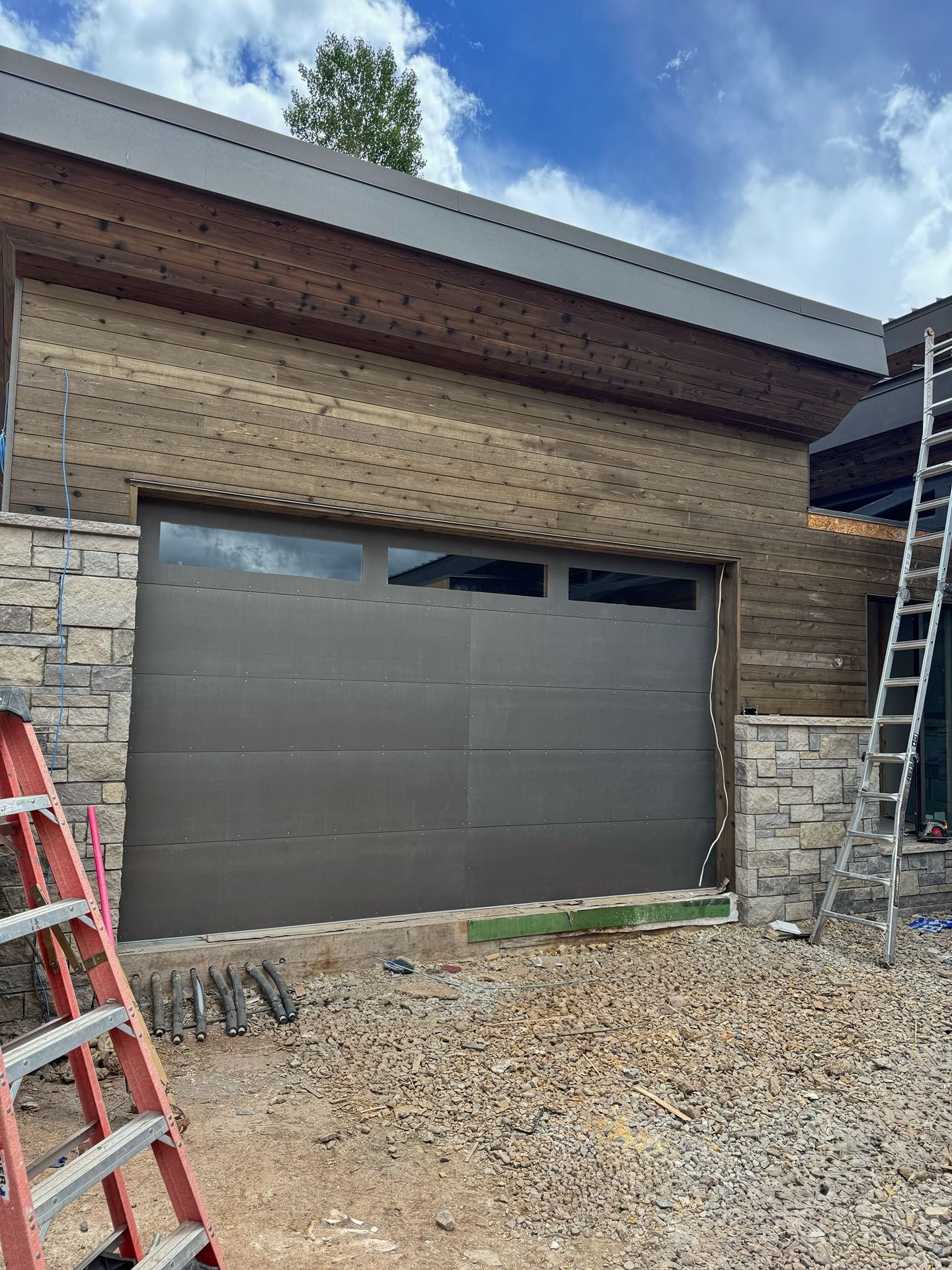 A modern garage with a dark gray door and rectangular windows, set against stone masonry and wooden siding under a blue sky.
