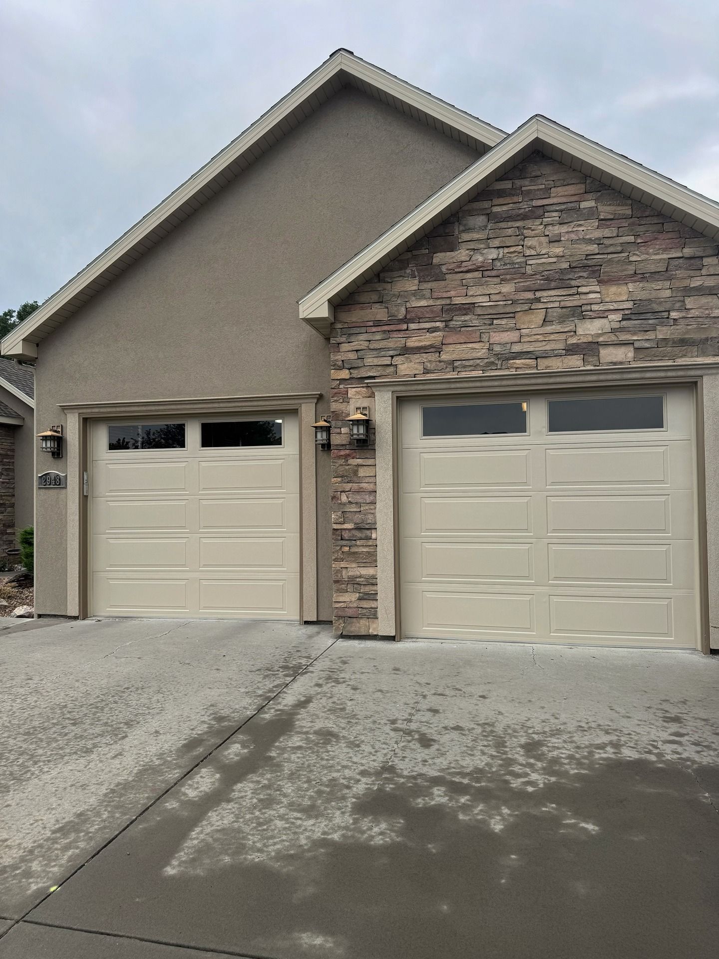 A tan, two-car garage with stone siding and light-colored doors under a peaked roof.