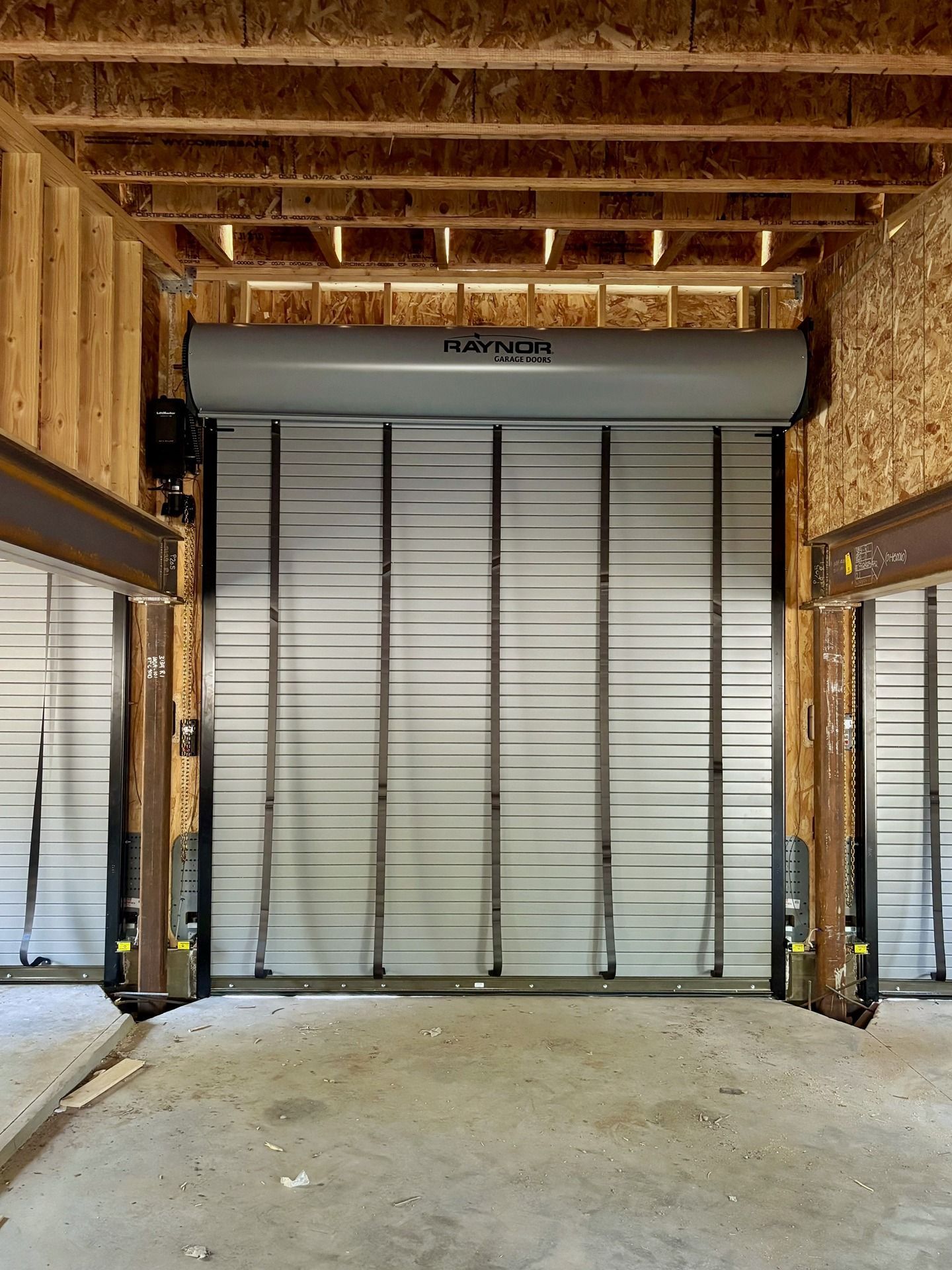 A grey, industrial roll-up door installed between wooden support beams in an unfinished building interior.