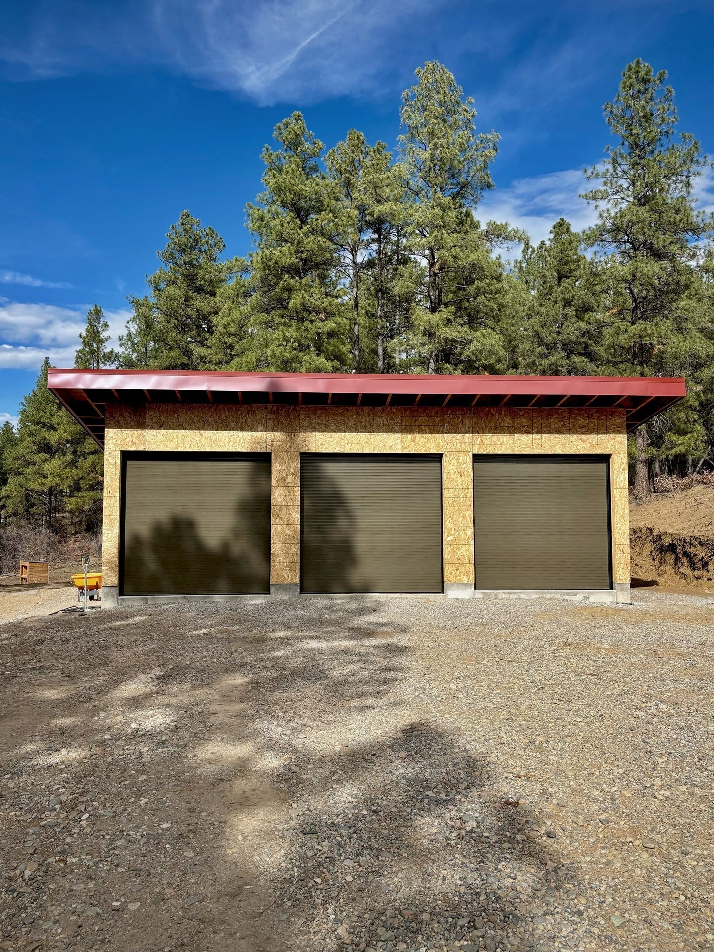 A newly built, single-story garage with three brown doors and oriented strand board siding sits before a forest.