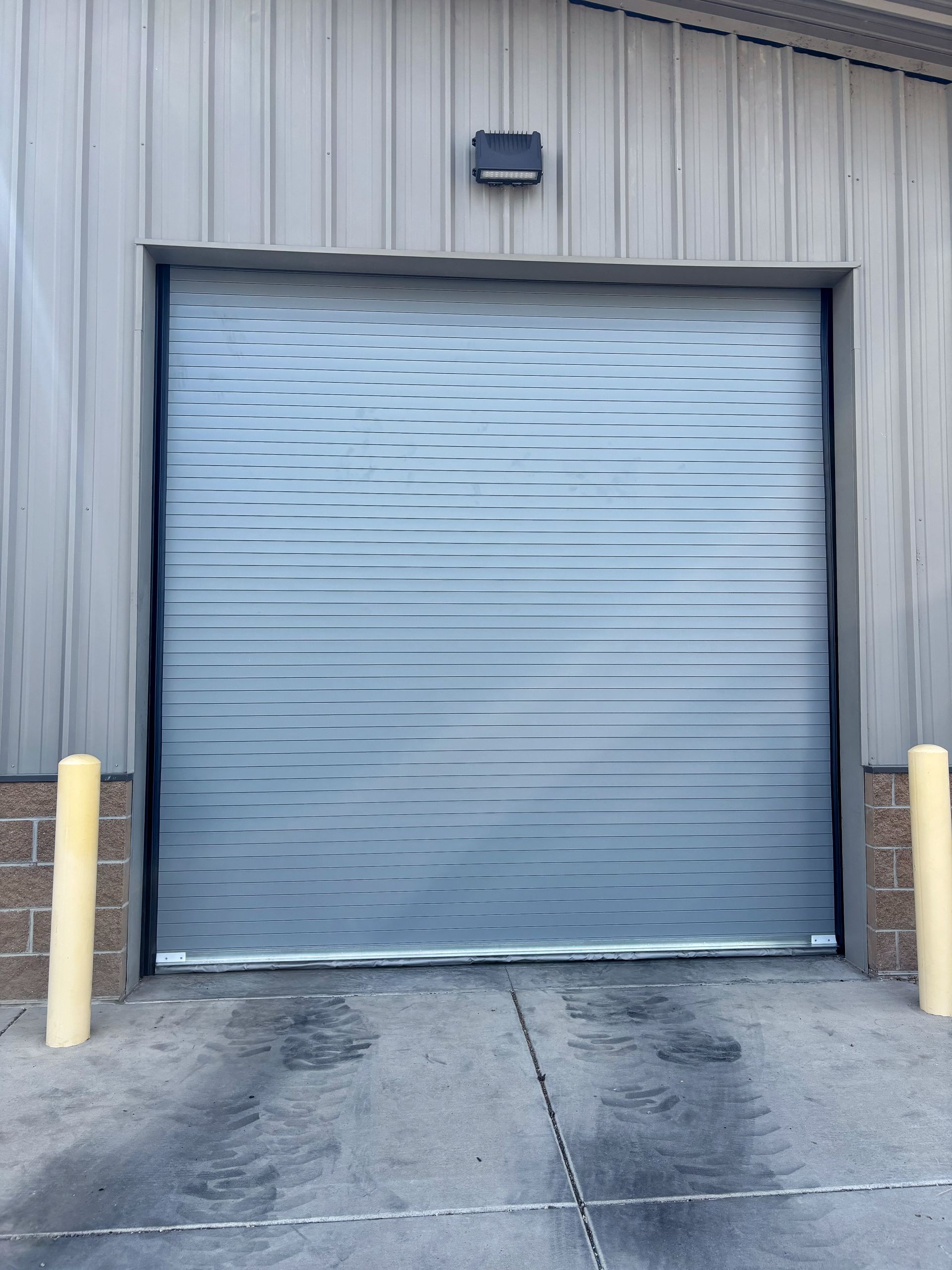 Closed gray roll-up garage door on a metal warehouse wall with concrete pavement in front