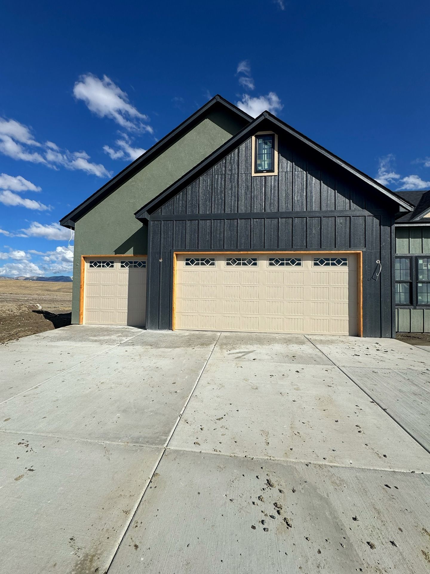 A house exterior featuring a dark board-and-batten garage, a green side wall, beige garage doors, and a concrete driveway.
