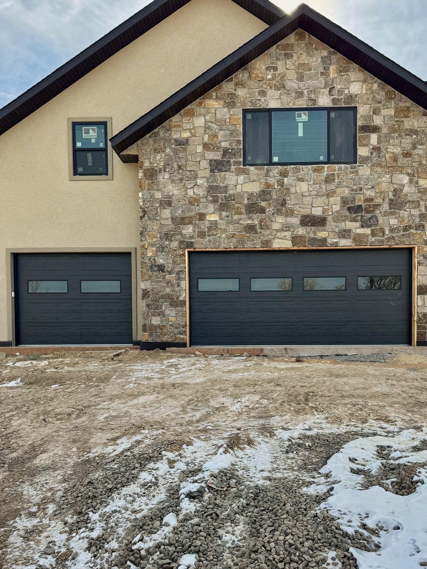 A tan stucco and stone house facade with two dark gray garage doors with horizontal windows under a partially cloudy sky.