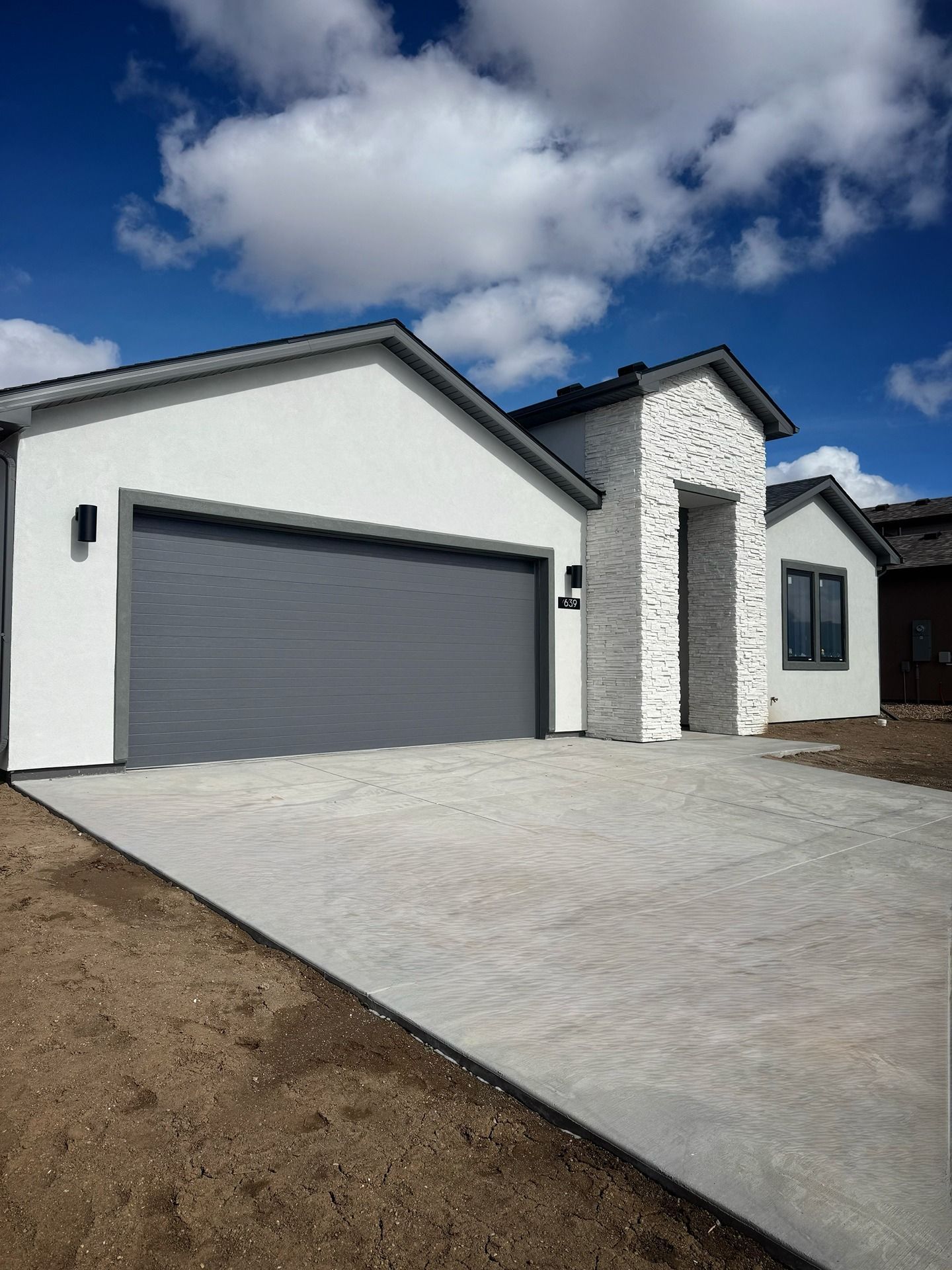 A modern, single-story house with white stucco walls, a grey garage door, and a white stone-faced entryway under a blue sky.