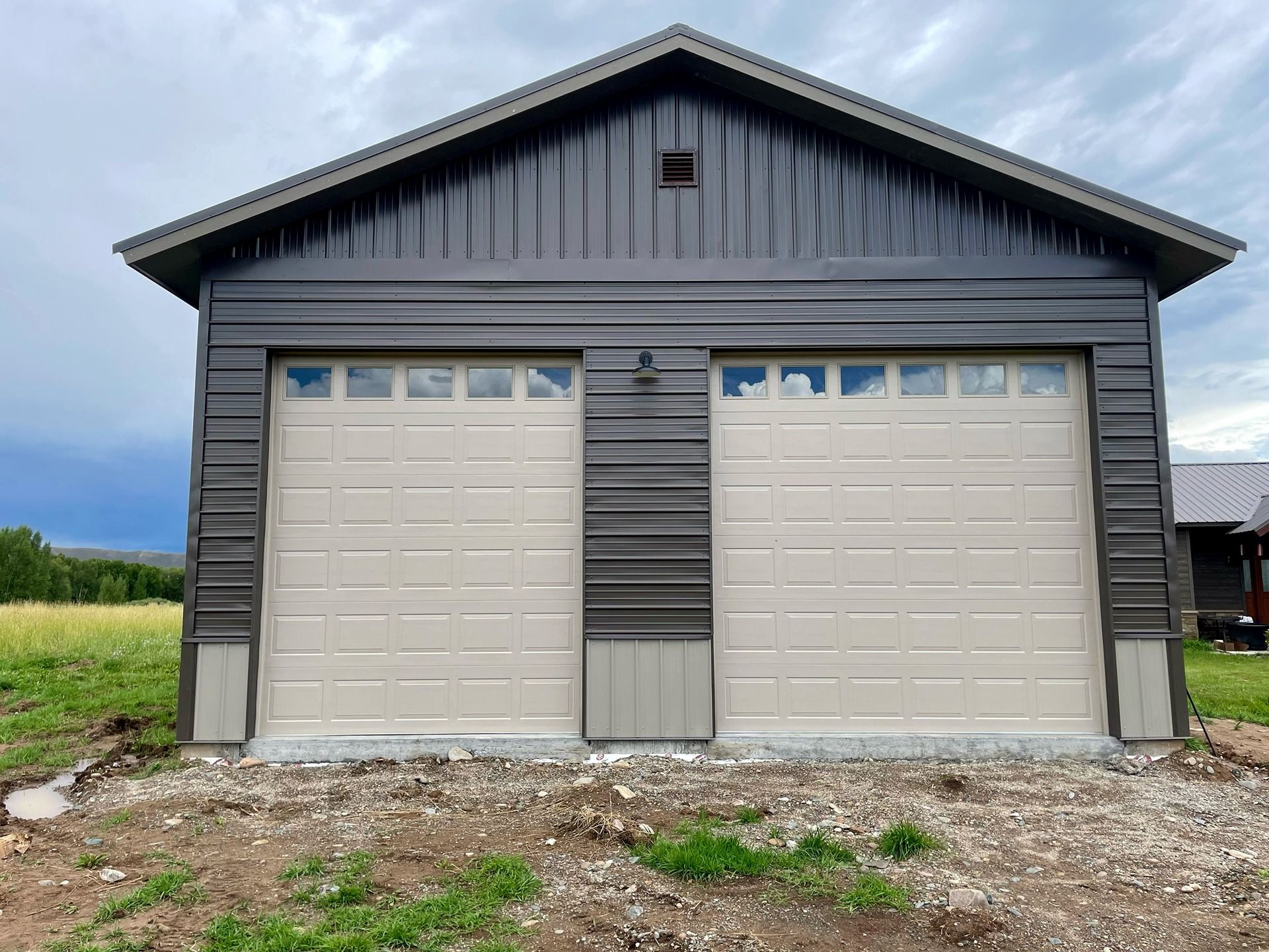 A detached two-car garage with charcoal metal siding and two beige garage doors in a grassy, rural setting.