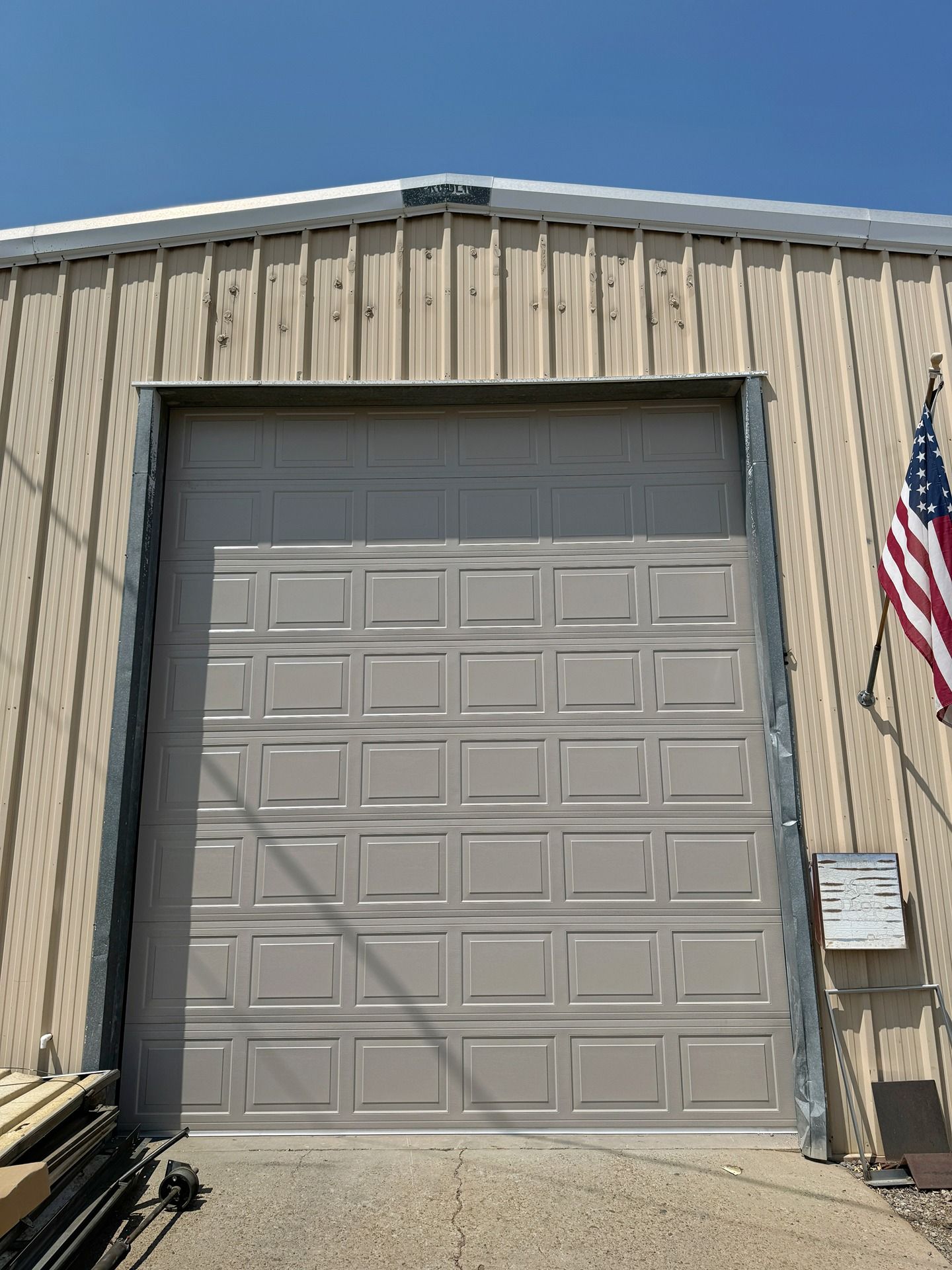 Tan metal building exterior featuring a large, grey sectional roll-up garage door with an American flag attached.