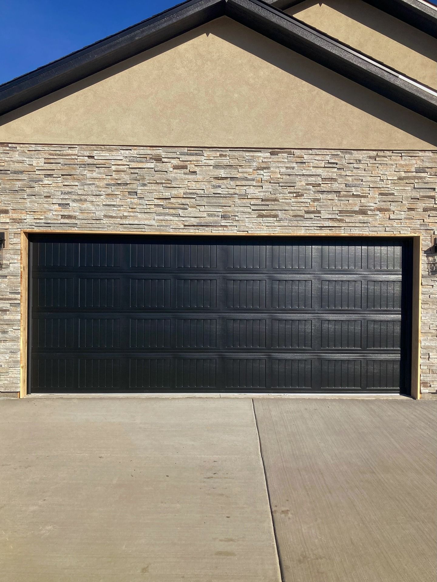 A black, rectangular garage door centered on a house exterior with stone and tan siding and a concrete driveway.