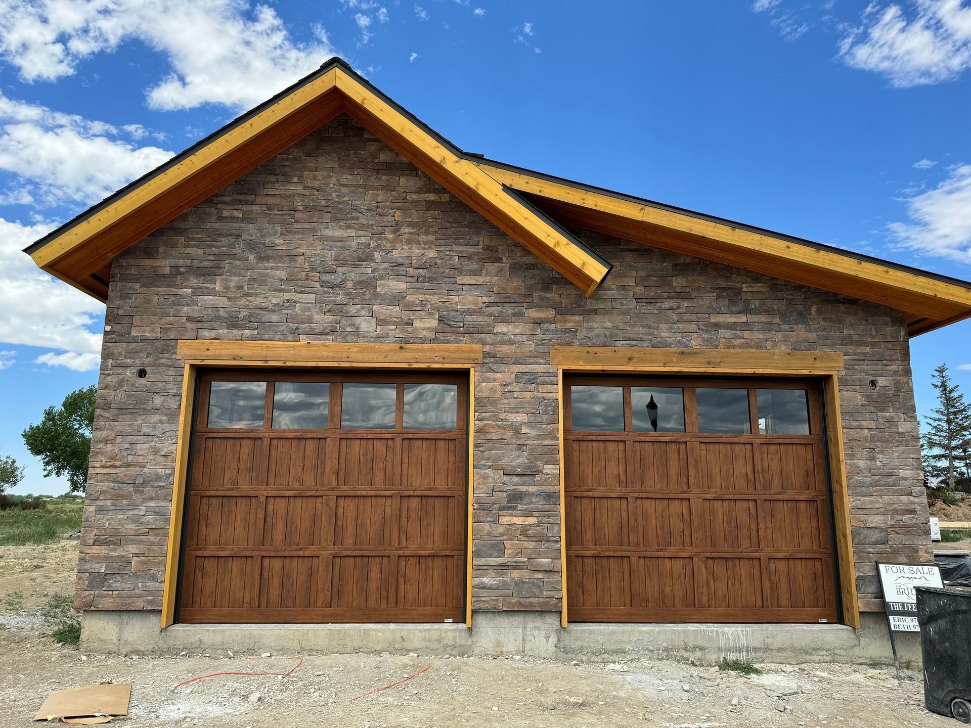 A modern detached garage with stone siding, two brown wood-look garage doors, and a natural wood trim roofline.
