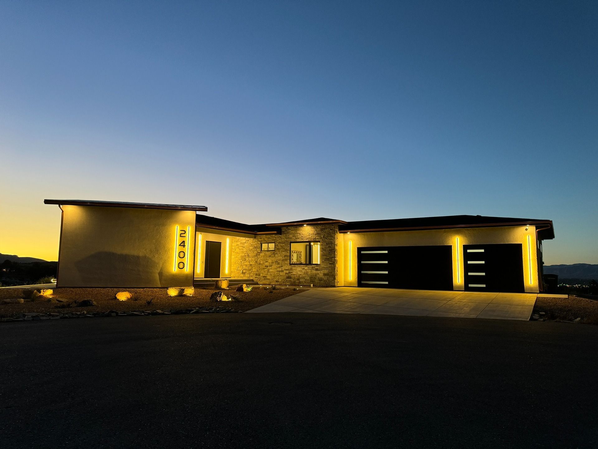 A modern, one-story house with illuminated address numbers and garage lights at twilight against a sunset sky.