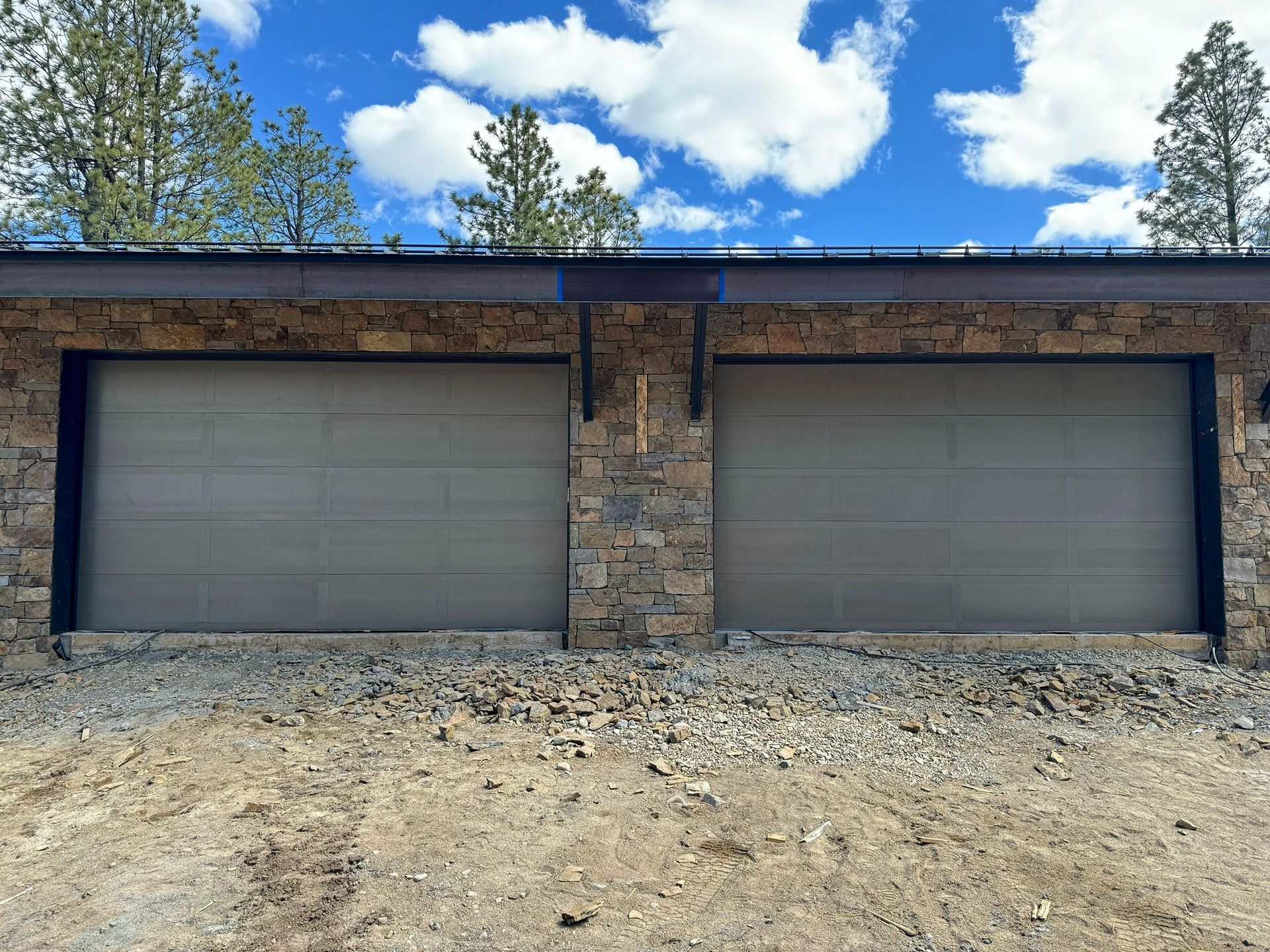 Two beige garage doors set into a rustic stone building under a blue, cloudy sky, with a gravel driveway in front.