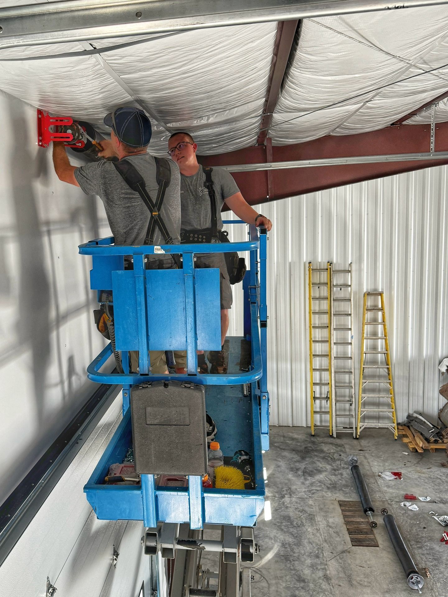 Two workers in a blue scissor lift install a red track onto a wall inside a building with a white insulated ceiling.
