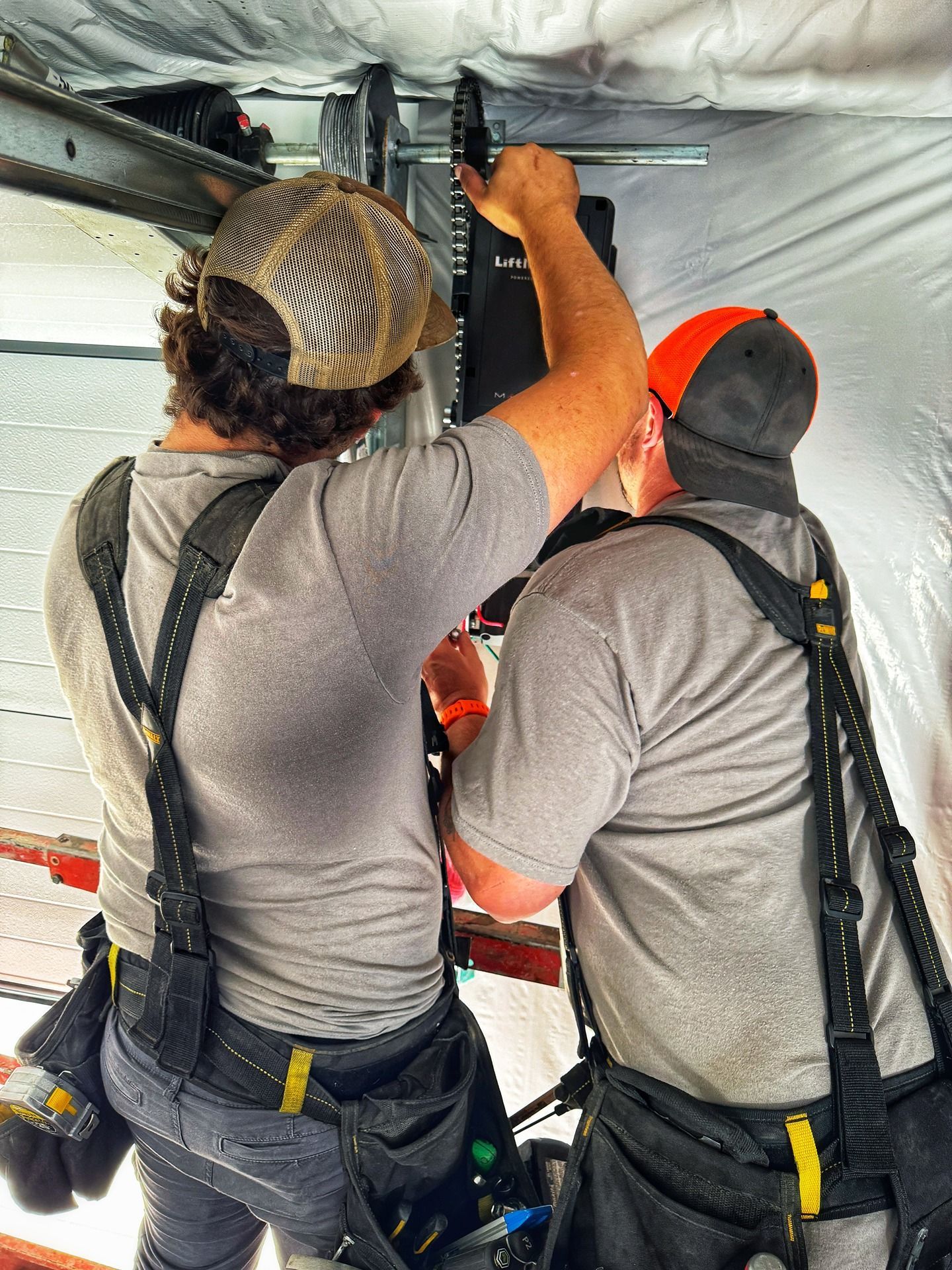 Two workers in grey shirts and tool belts installing a garage door motor mechanism on a ceiling track.