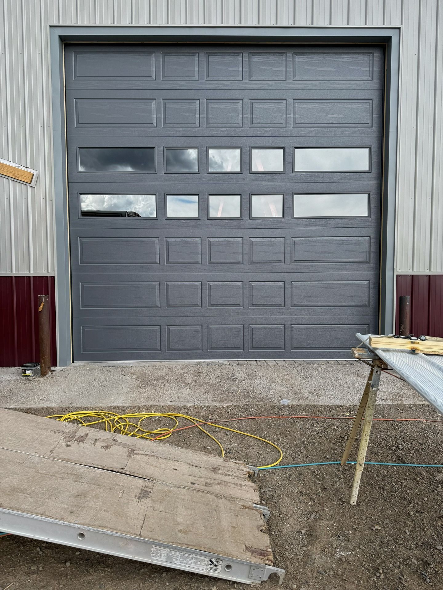 A dark gray metal garage door with horizontal windows on a metal building, with construction equipment in the foreground.
