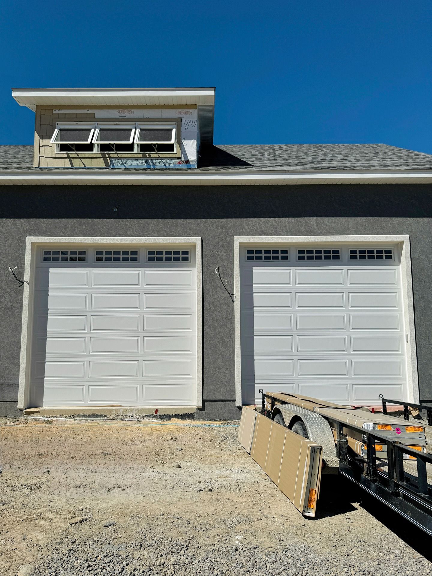 Two white garage doors on a grey building under a clear blue sky, with a trailer parked in front.