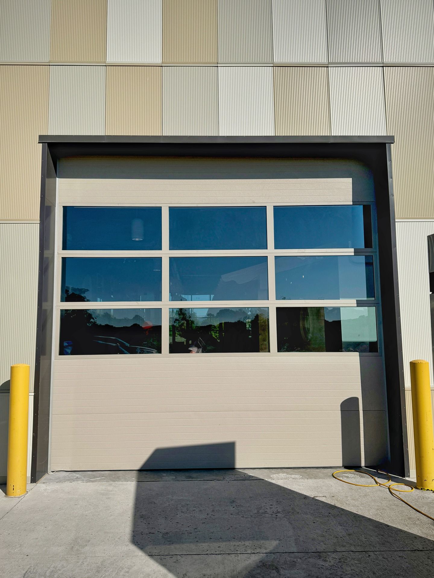 A beige industrial bay door with a horizontal row of three rectangular glass windows, flanked by two yellow bollards.