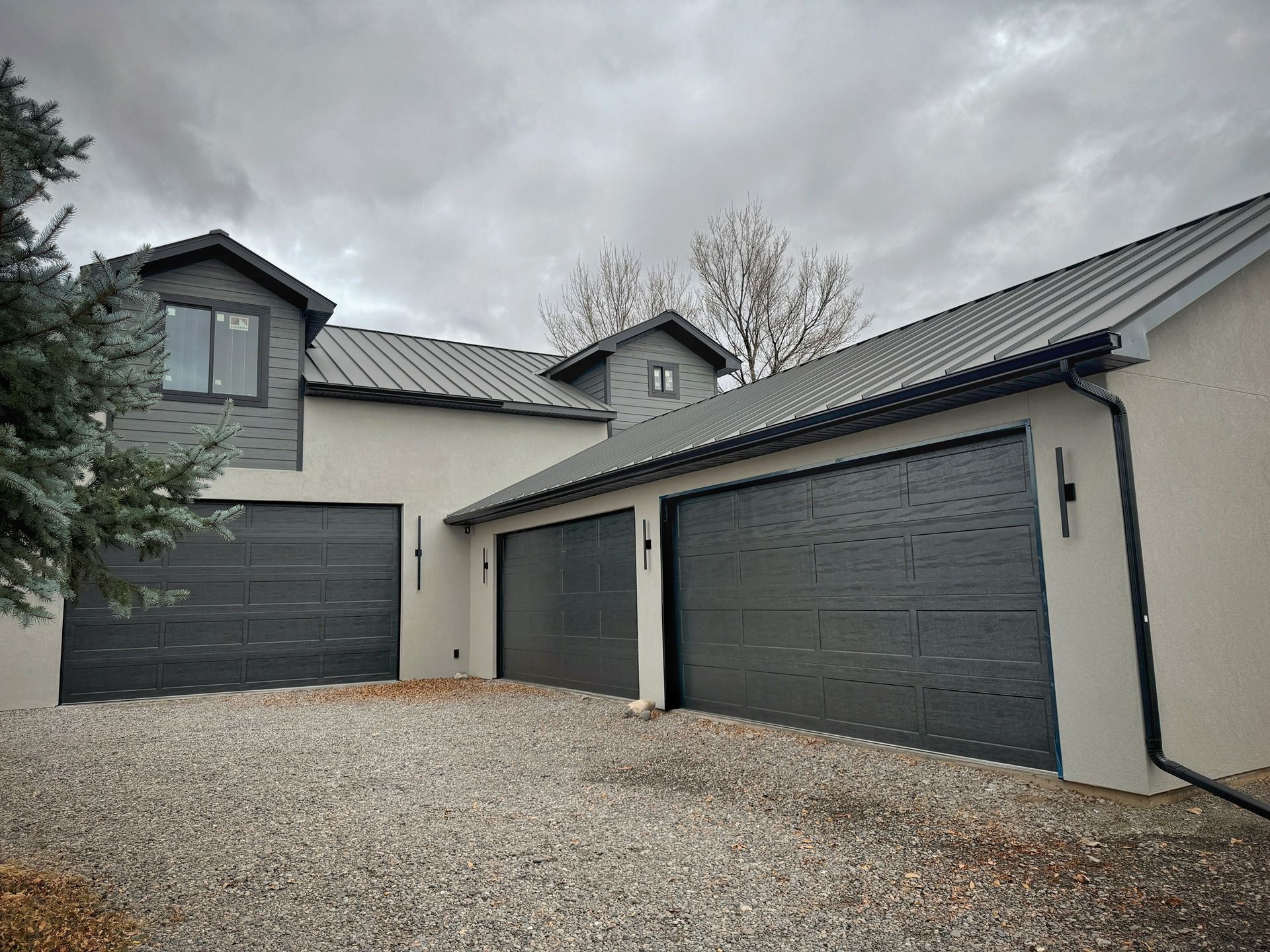 A modern house with light beige stucco walls, dark grey metal roofs, and three matching dark grey garage doors.