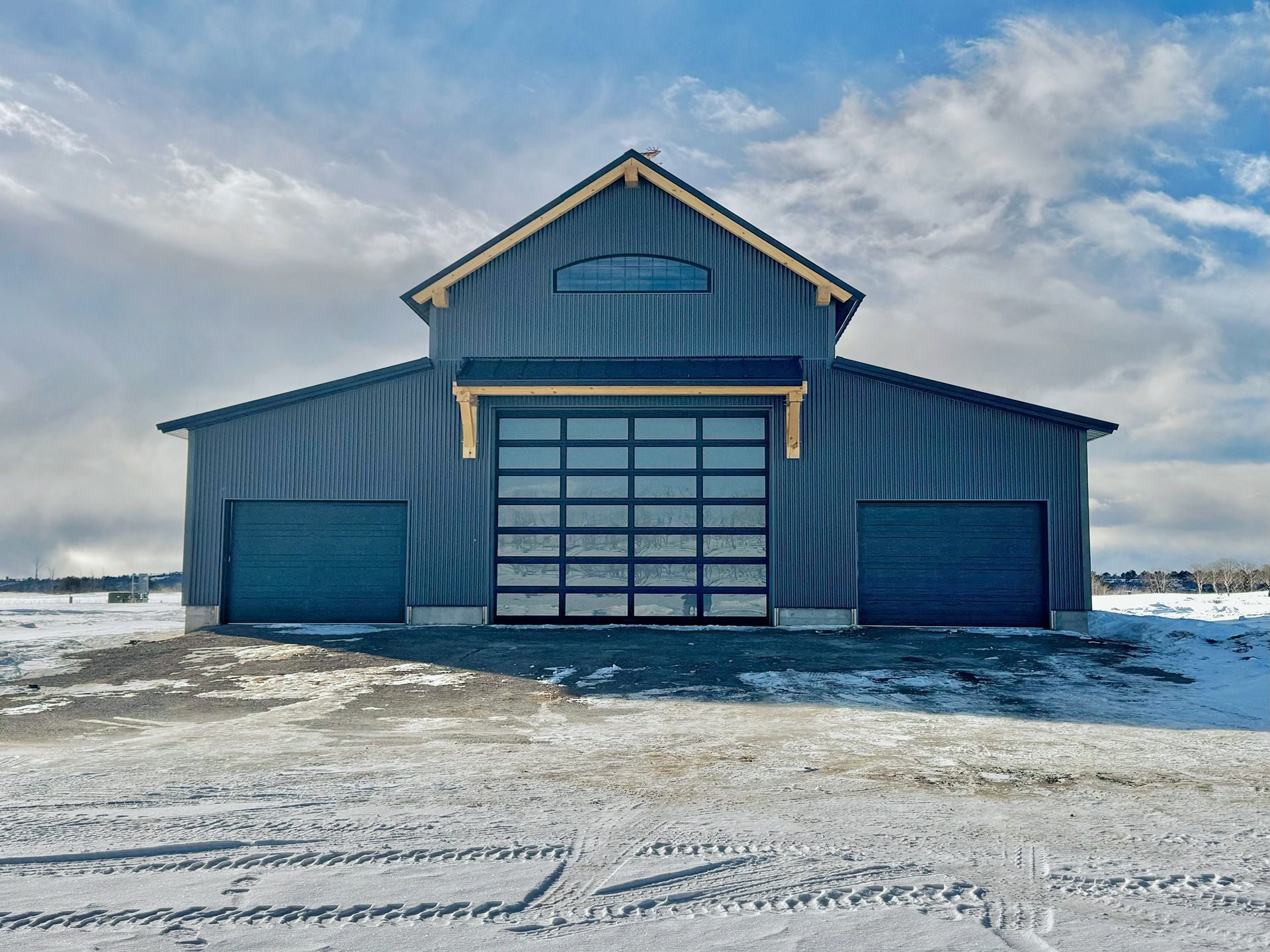 A dark blue barn with a central glass garage door and two side doors, set in a snowy landscape under a cloudy sky.