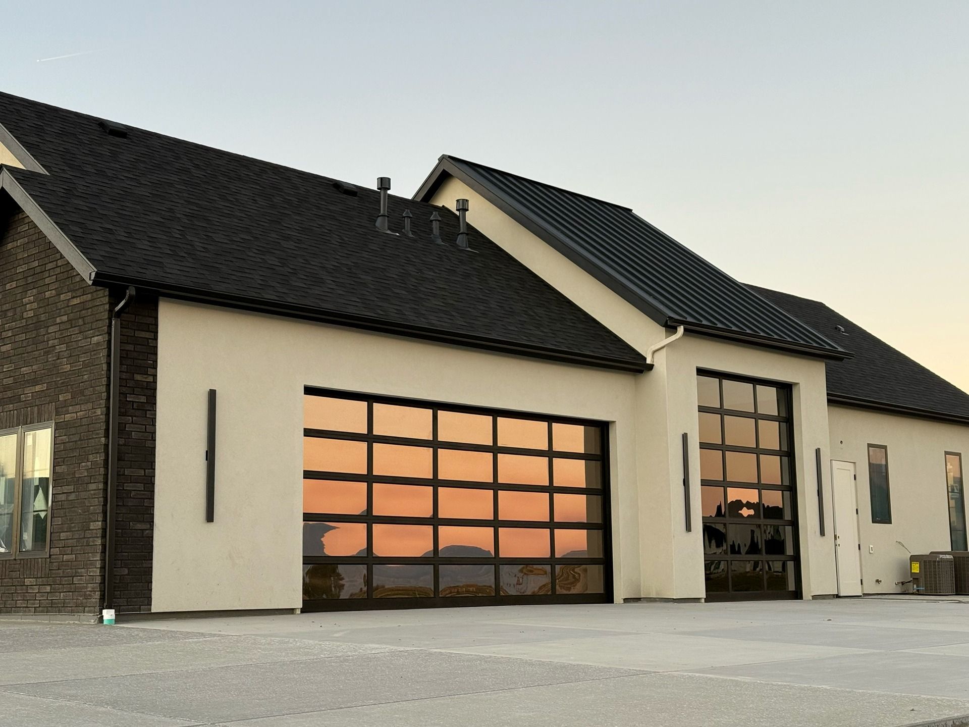 Modern single-story home with a tan stucco exterior, black roof, and two large glass-paneled garage doors reflecting sunset.
