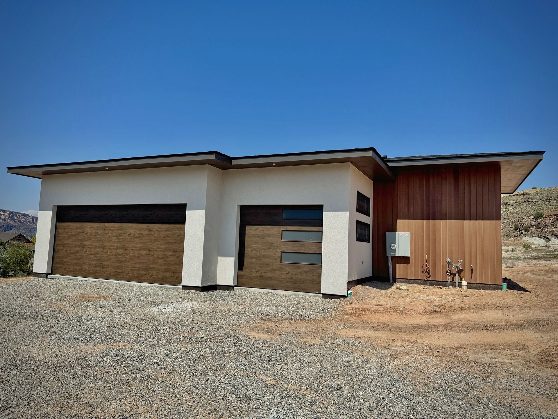 Modern single-story home with light stucco, wood paneling, and two garage doors on a rocky, desert lot under a blue sky.