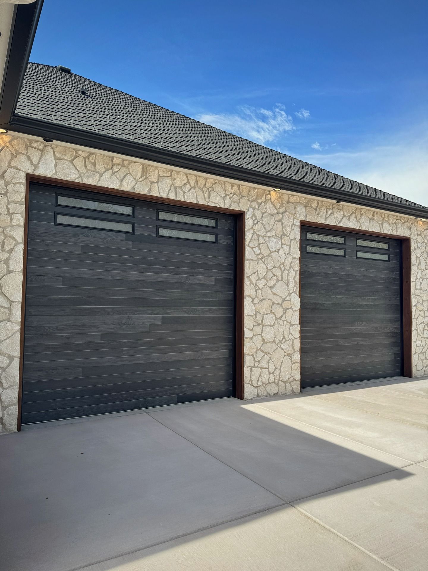 Two modern dark gray garage doors with small rectangular windows, set into a light-colored stone exterior wall.