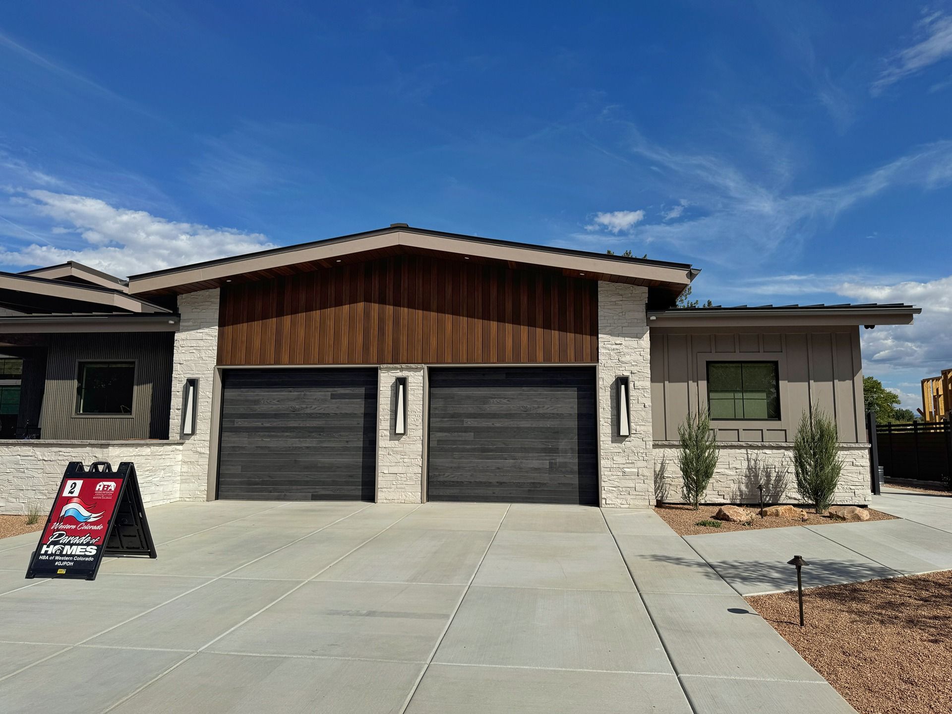 Modern suburban home exterior featuring two dark garage doors, stone accents, and wood siding under a clear blue sky.