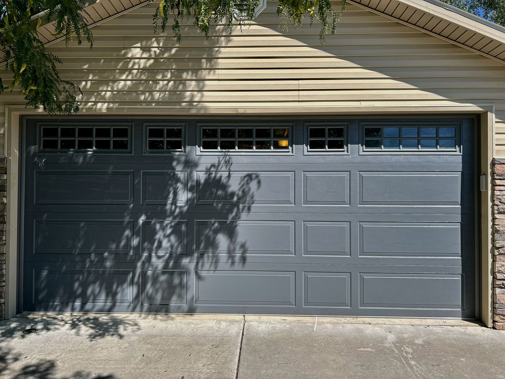 A modern, dark grey, single-car garage door with decorative rectangular windows at the top, set into a beige home exterior.
