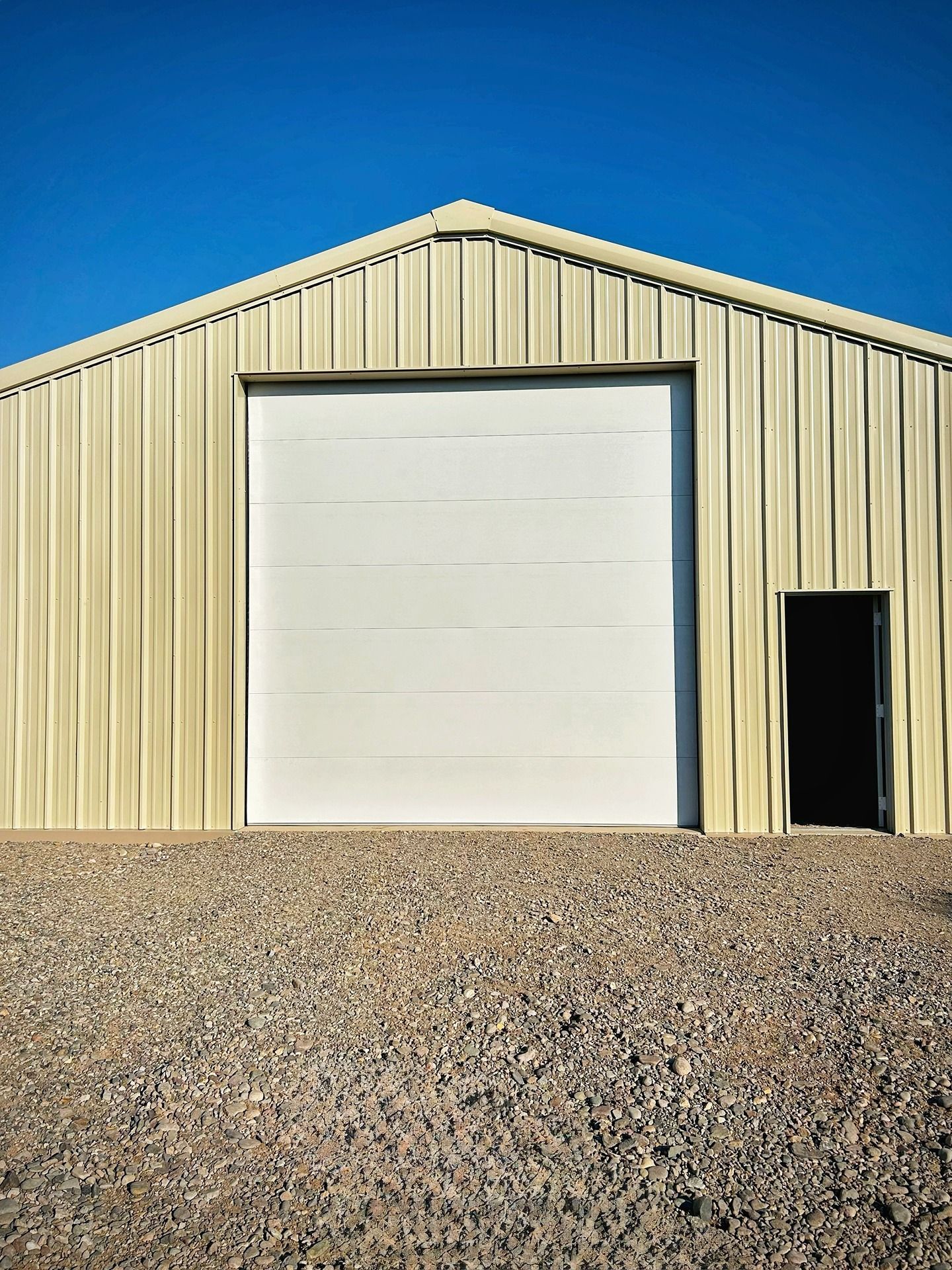 A cream-colored metal shed with a large white roll-up door and a smaller side entry door, set on a gravel lot under blue sky.