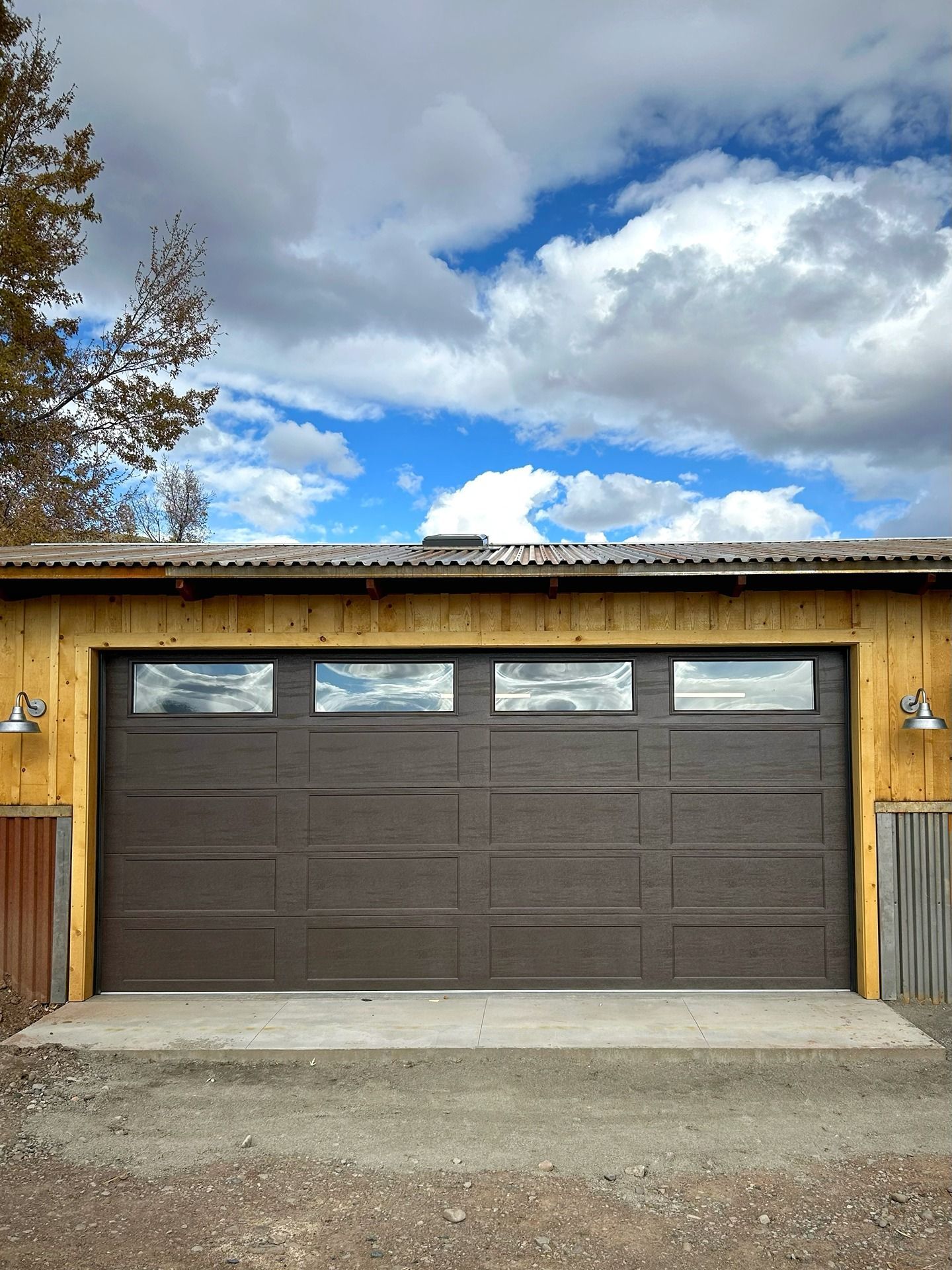 A brown garage door with four top window panels set in a light wood-paneled building under a bright, cloudy blue sky.