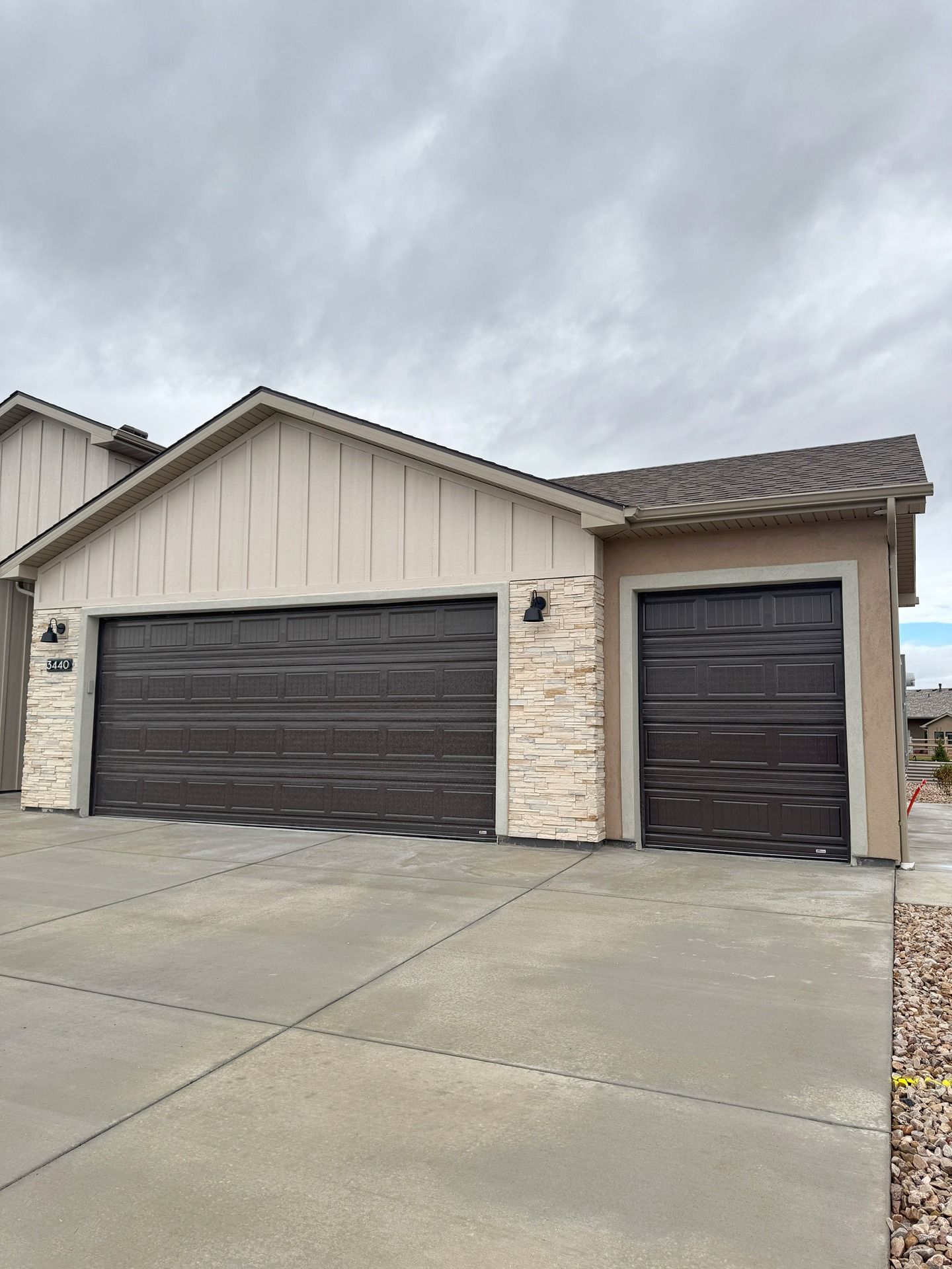 A modern suburban house exterior featuring a beige vertical-sided gable, stone accents, and two dark brown garage doors.