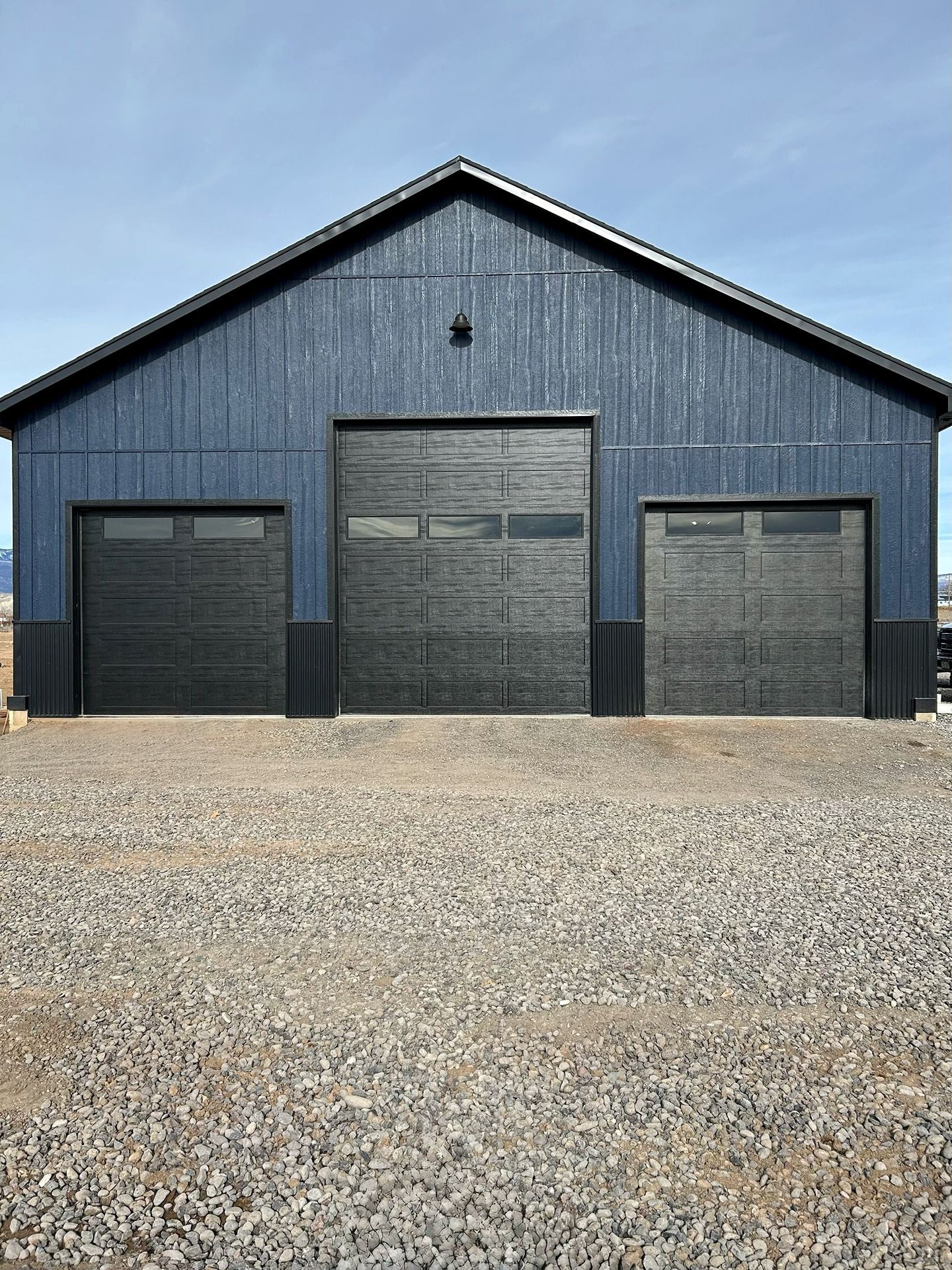 A blue, gabled-roof barn featuring one large central garage door and two smaller side garage doors on a gravel lot.