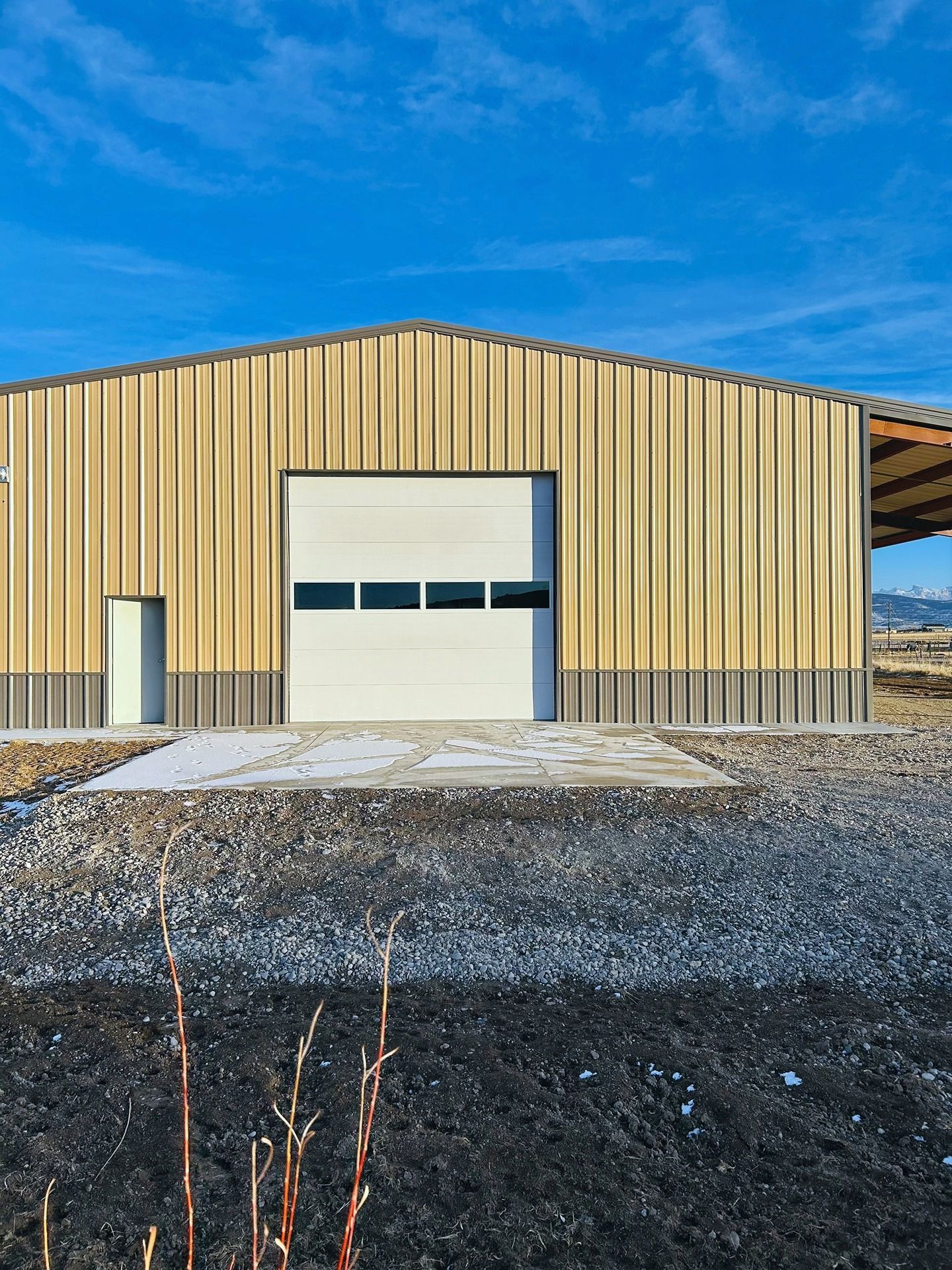 A modern metal building with tan and gray siding, a white garage door, and a man door under a bright blue sky.