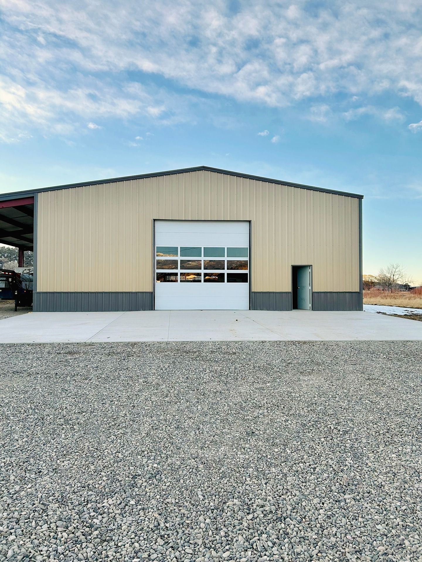 A tan and grey metal workshop building with a white glass-paneled garage door and side entrance, set above a gravel lot.
