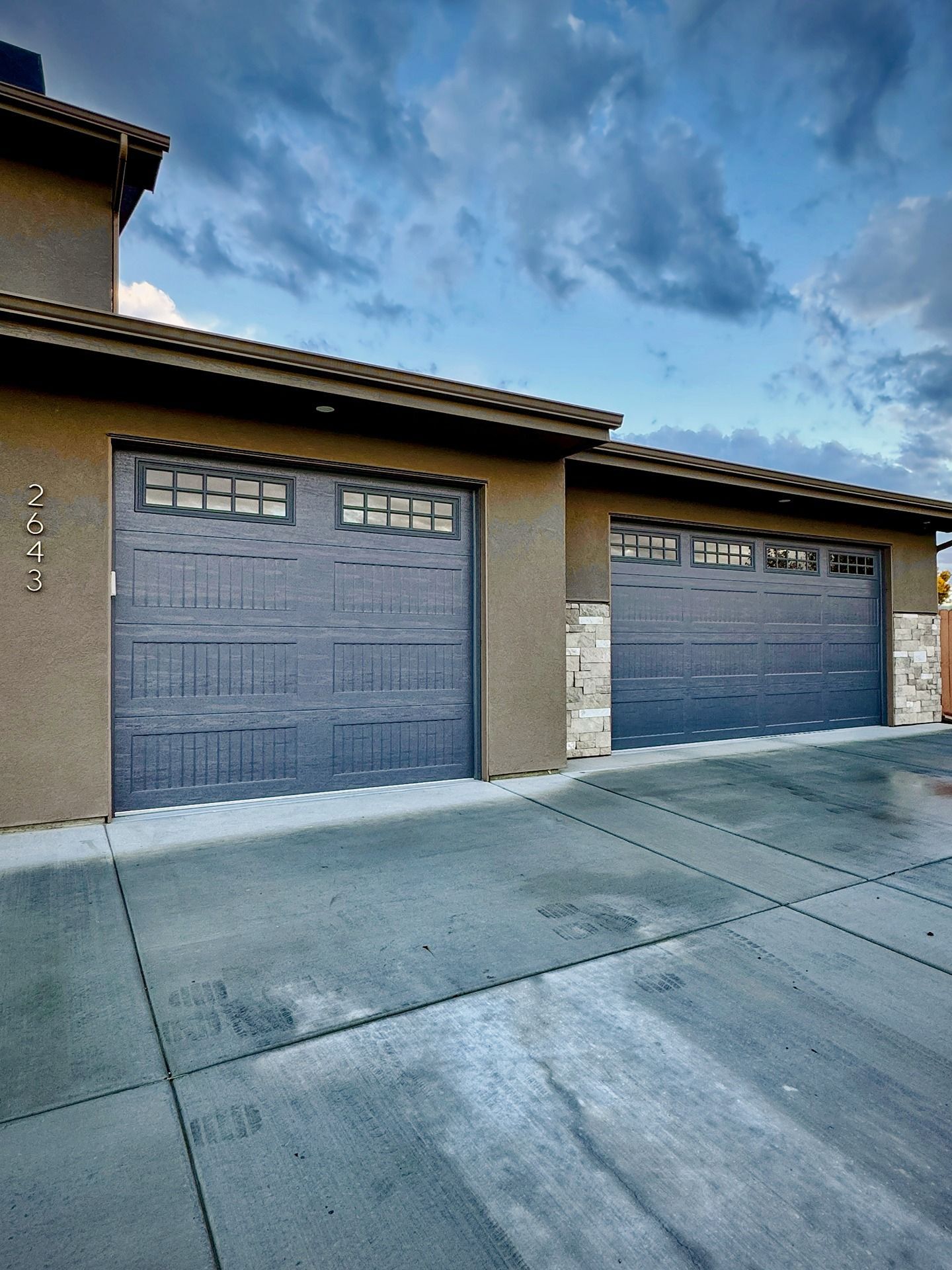 A two-car garage with gray paneled doors, stone accents, and a concrete driveway under a cloudy blue sky.