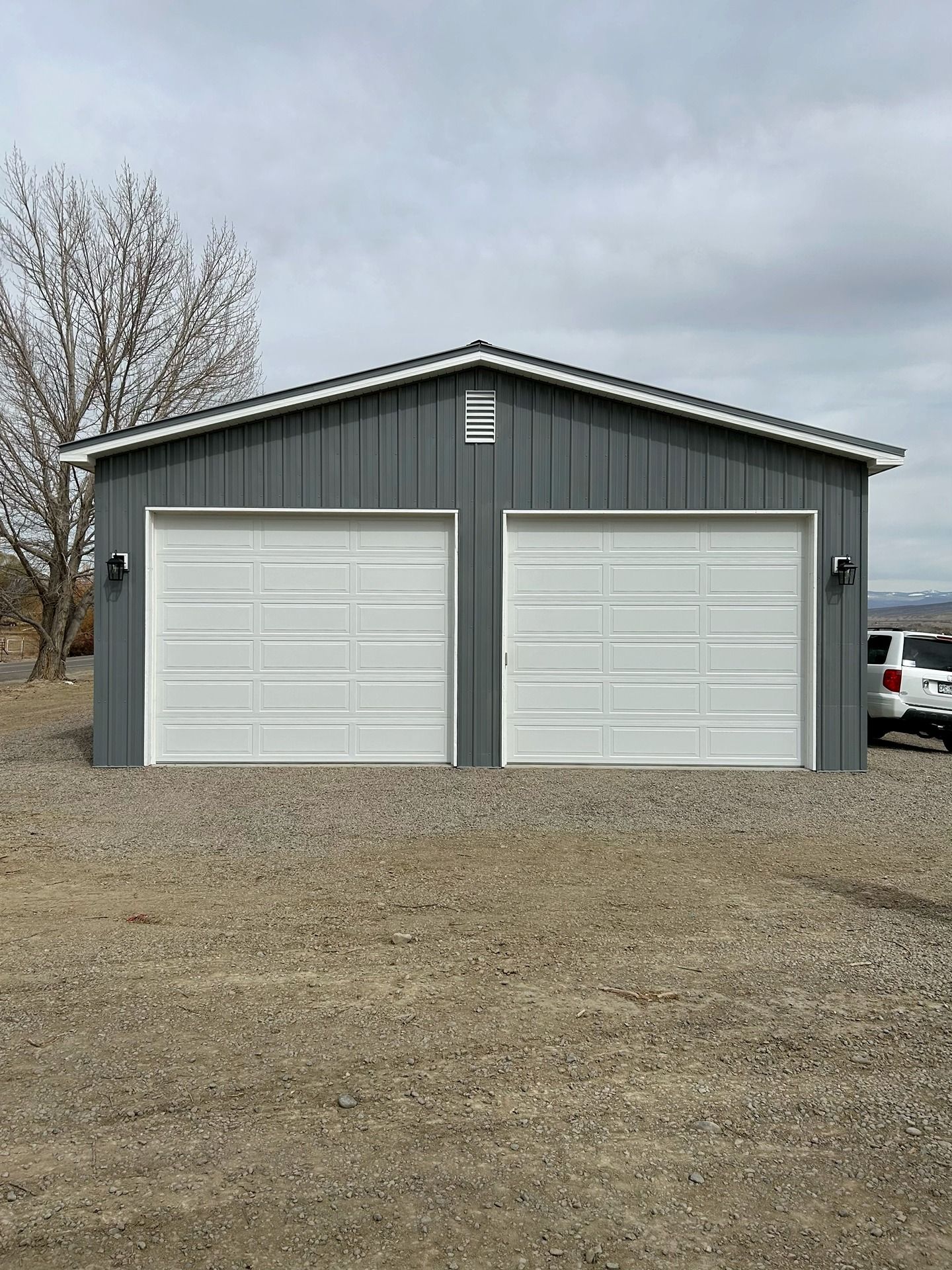A gray metal-sided garage with two white roll-up doors, set on a gravel lot under a cloudy sky.
