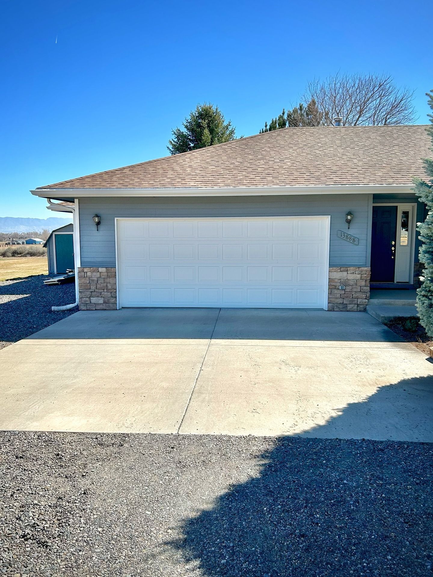 A gray house exterior with a two-car white garage door, stone accents, and a concrete driveway under a clear blue sky.