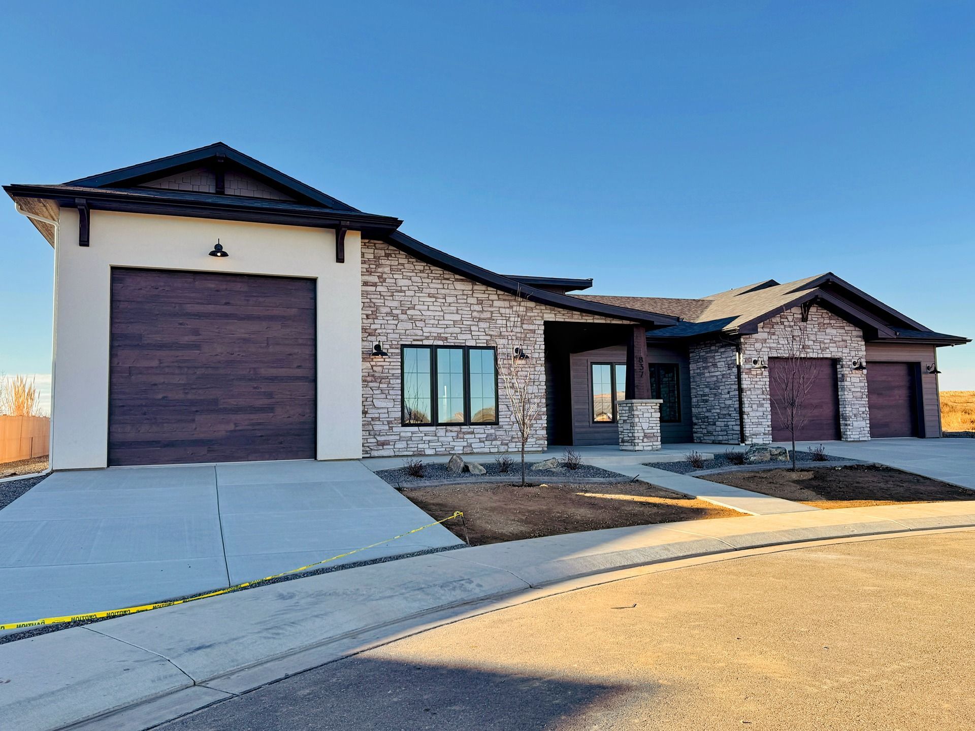Single-story modern suburban house with a three-car garage, light stone siding, and dark trim against a clear blue sky.