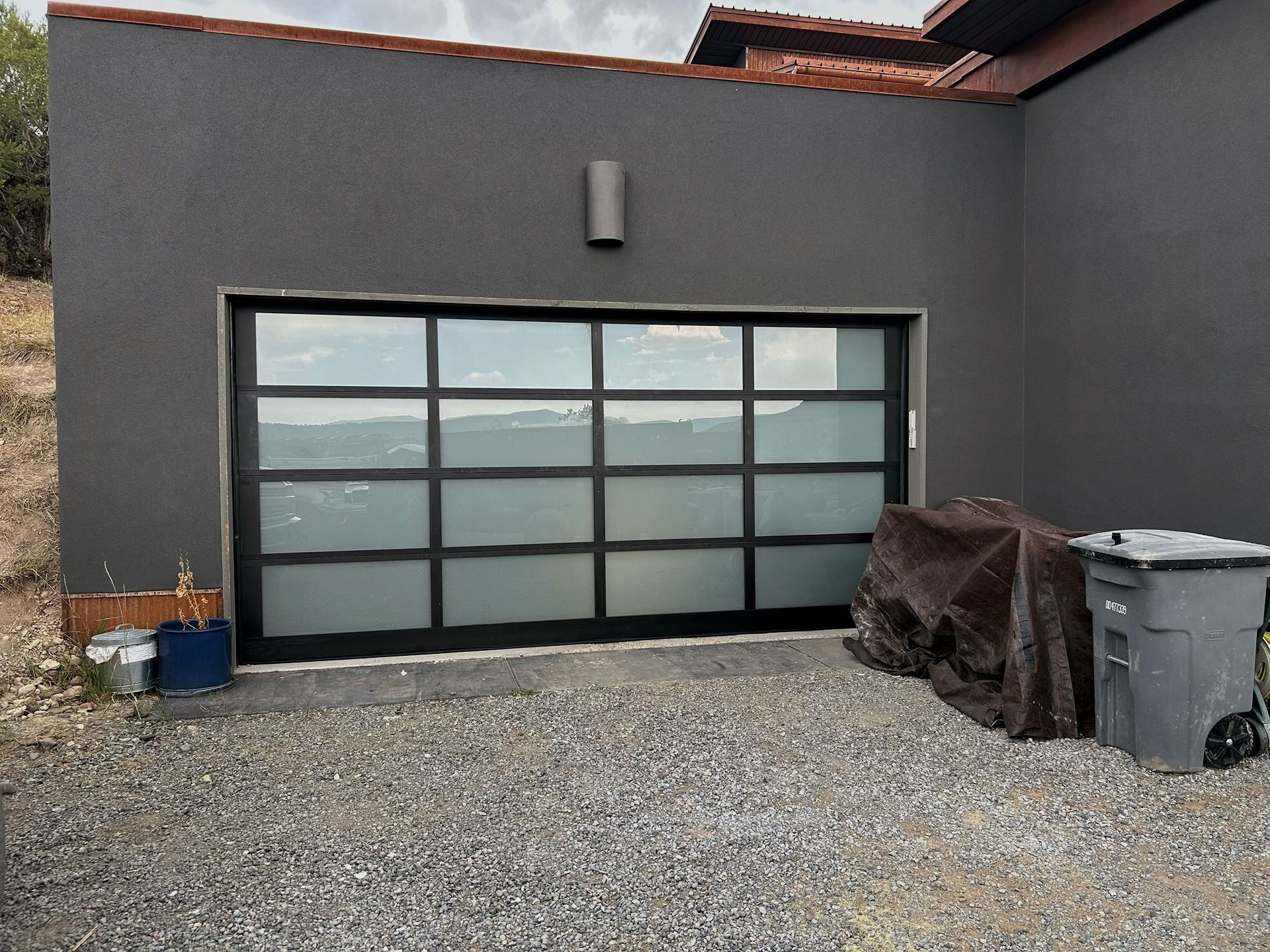 A modern garage with a dark gray facade and a large, multi-panel glass garage door sits on a gravel driveway.