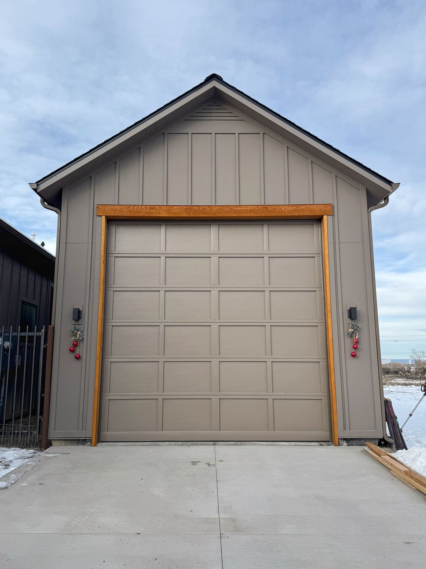 A tan-colored garage with a panelled door framed in wood, set against a cloudy sky with patches of snow on the ground.