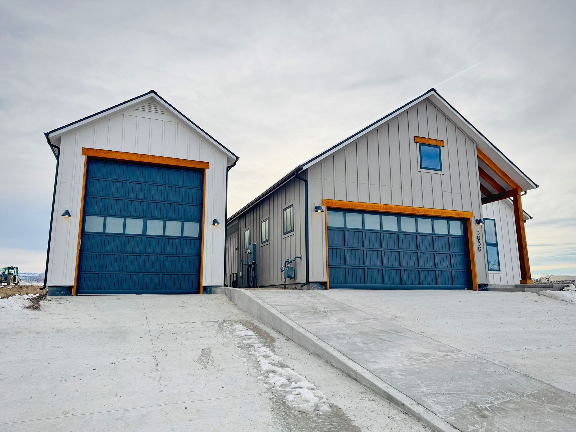 Two modern white buildings with blue garage doors and wood trim, situated on a snowy lot under a cloudy sky.