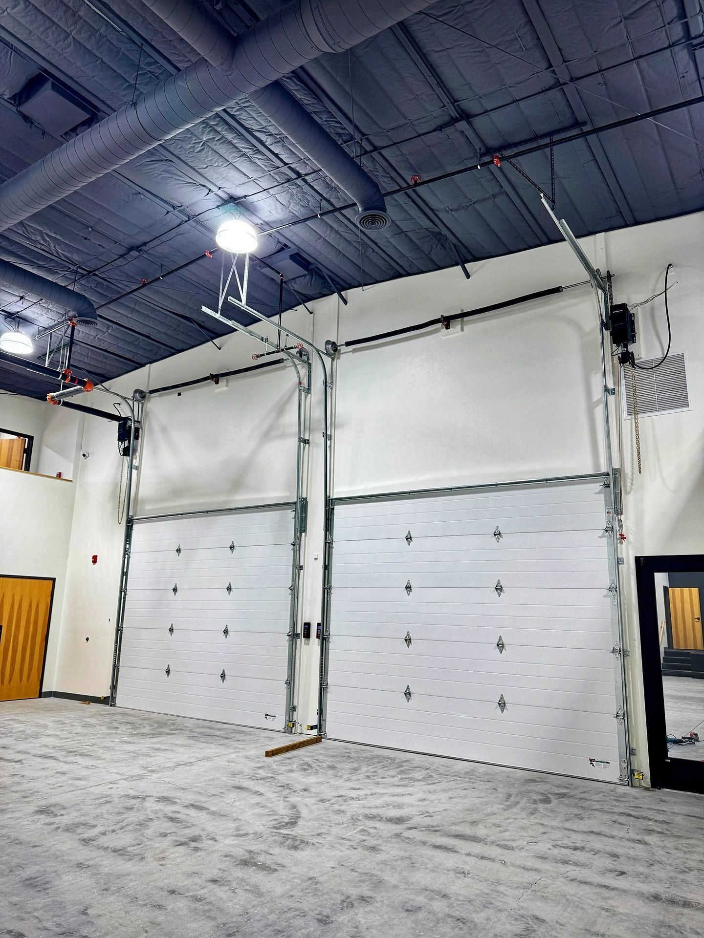 Two large, white industrial roll-up doors inside a spacious warehouse with an exposed black ceiling and concrete floor.