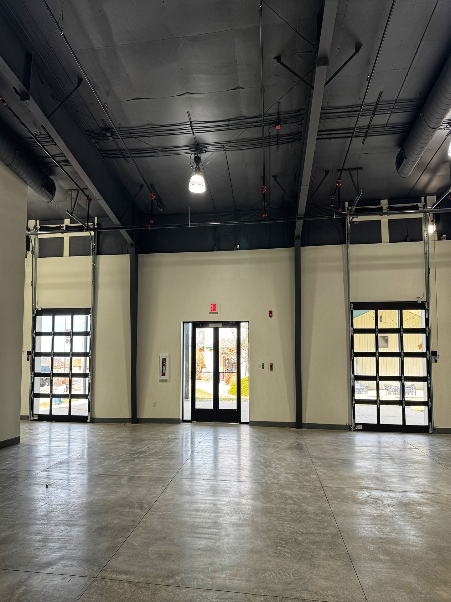 An empty industrial space with polished concrete floors, black ceiling, and three glass-paned doors leading outdoors.