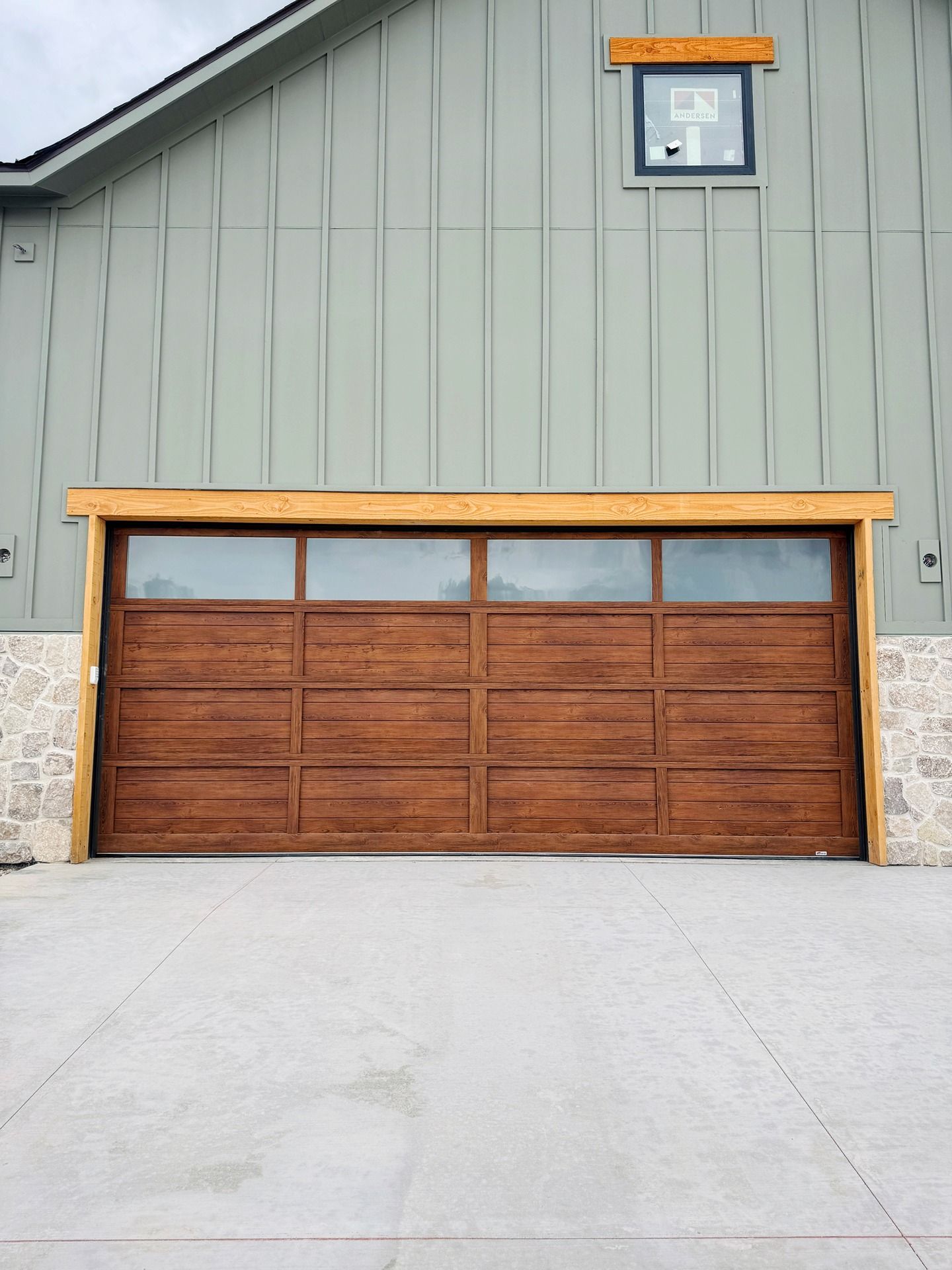 A dark wood garage door with a row of top windows, set in a light green barn-style building with stone accents.