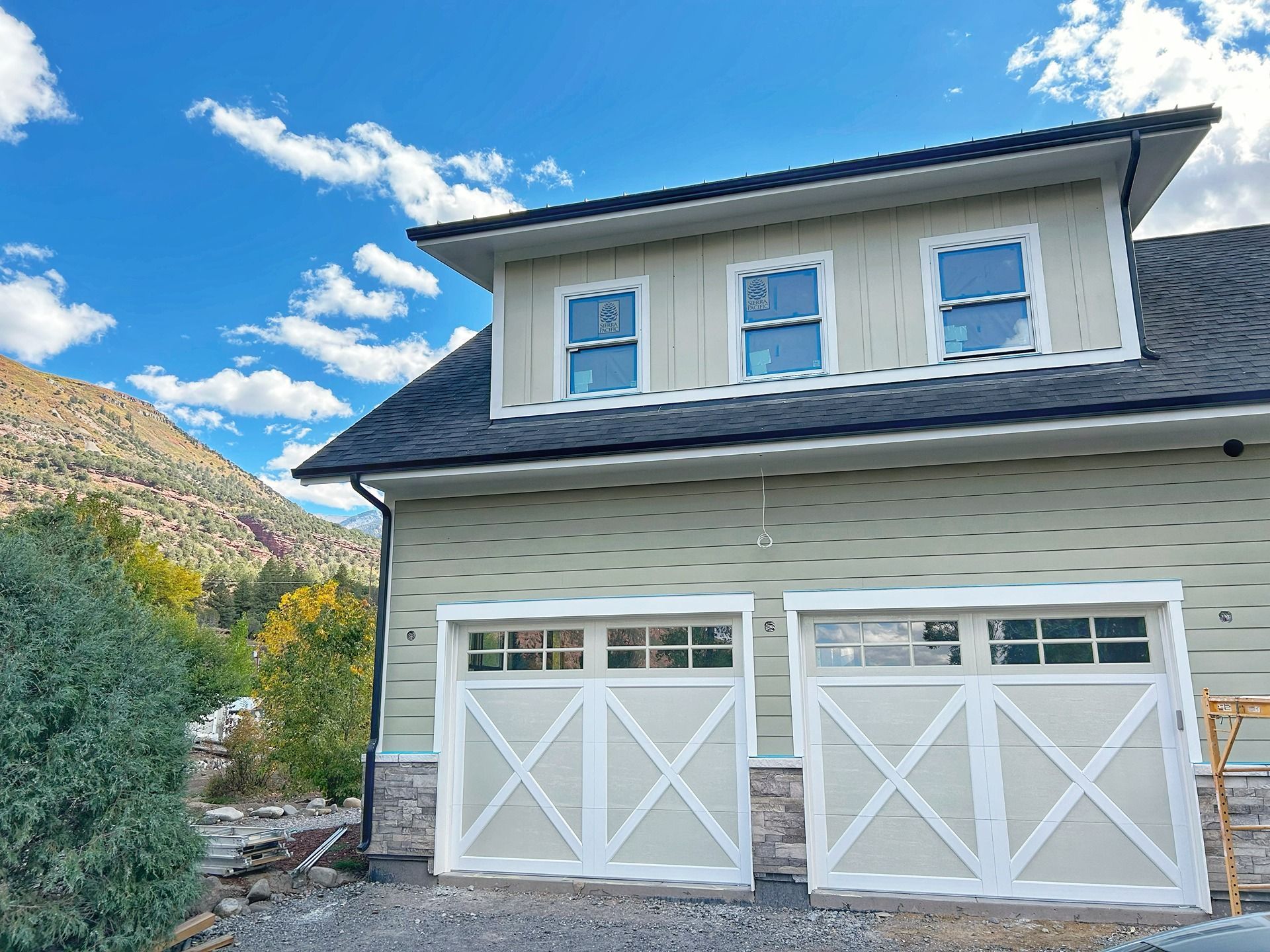 A light green, two-story garage with two white carriage-style doors, a dormer window, and stone accents against a mountain.